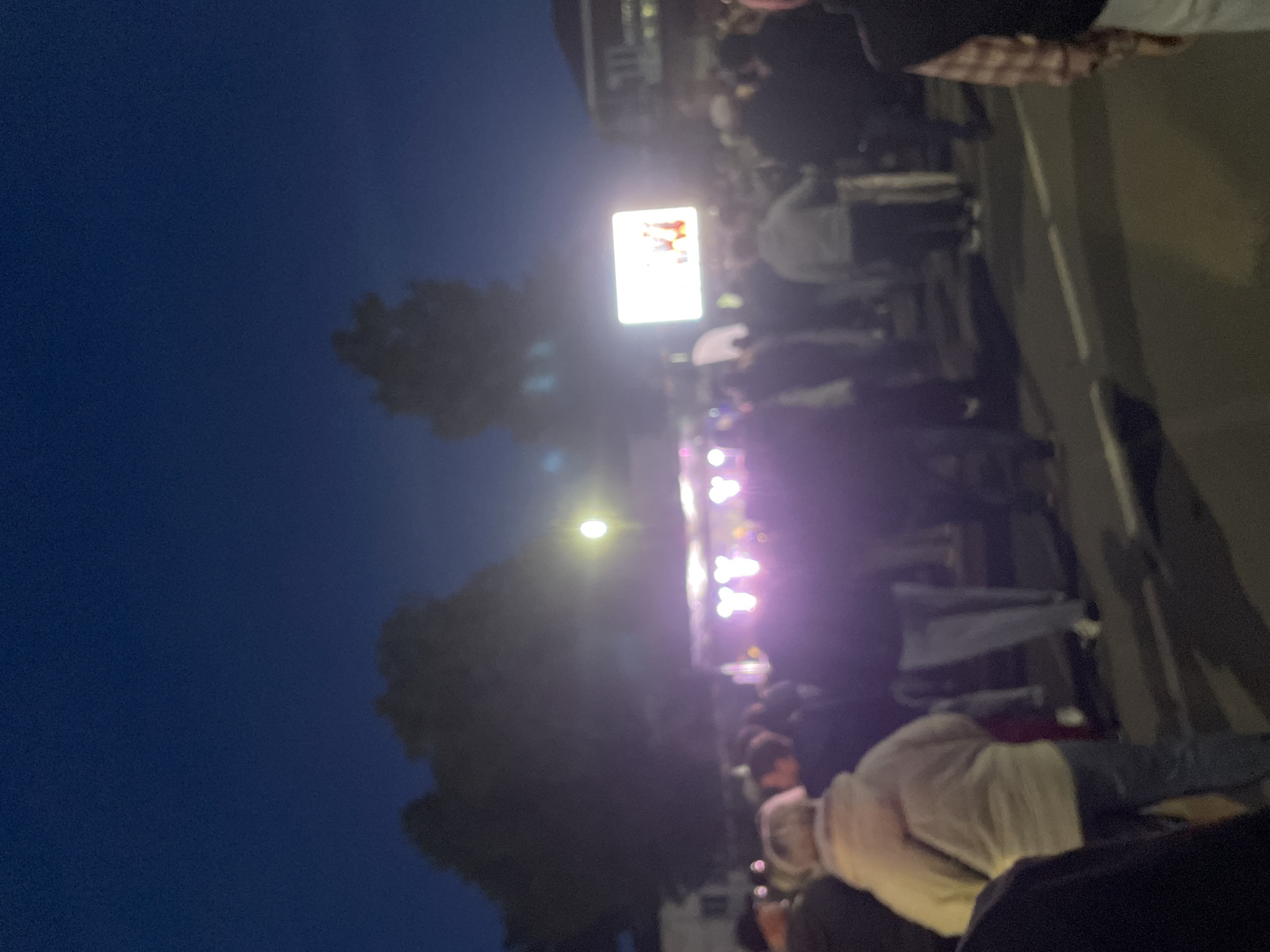 At night in Красные Баки, Russia, a crowd has gathered. A brightly lit screen is visible above the crowd. Stage lights are also present. Trees are seen to the left.
