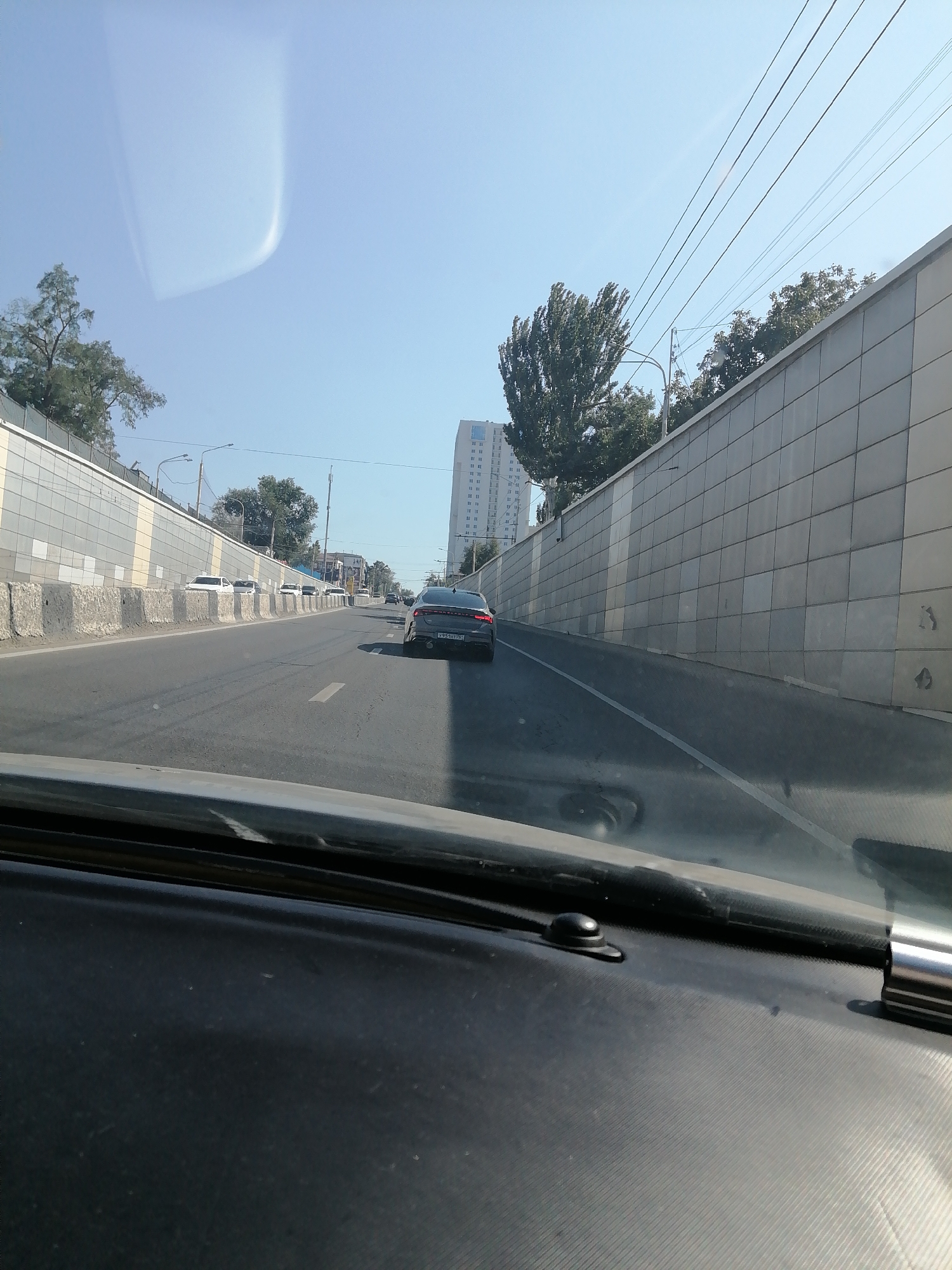 The image captures a view from inside a vehicle traveling on a multi-lane road, likely a highway or a major city thoroughfare. The road has distinct lane markings and is bordered by concrete barriers and retaining walls. The retaining wall on the right is constructed with large, square tiles and extends into the distance. On the left, a similar retaining wall is visible, with a fence on top of it and trees beyond.
Several cars are present on the road. In the foreground, the dashboard of the vehicle is visible, along with part of the windshield. Further down the road, a dark-colored sedan is driving in the right lane, its license plate partially obscured. Other vehicles are also seen in various lanes ahead.
In the background, a tall, multi-story building with numerous windows is situated on the right side of the road. The sky is clear and blue, with some subtle cloud formations that appear to be reflections on the windshield. The overall scene suggests a typical urban driving environment on a sunny day. While specific landmarks for Rostov-on-Don are not immediately apparent in this general view, the architectural style of the retaining walls and the presence of tall buildings are consistent with an urban setting in Russia.