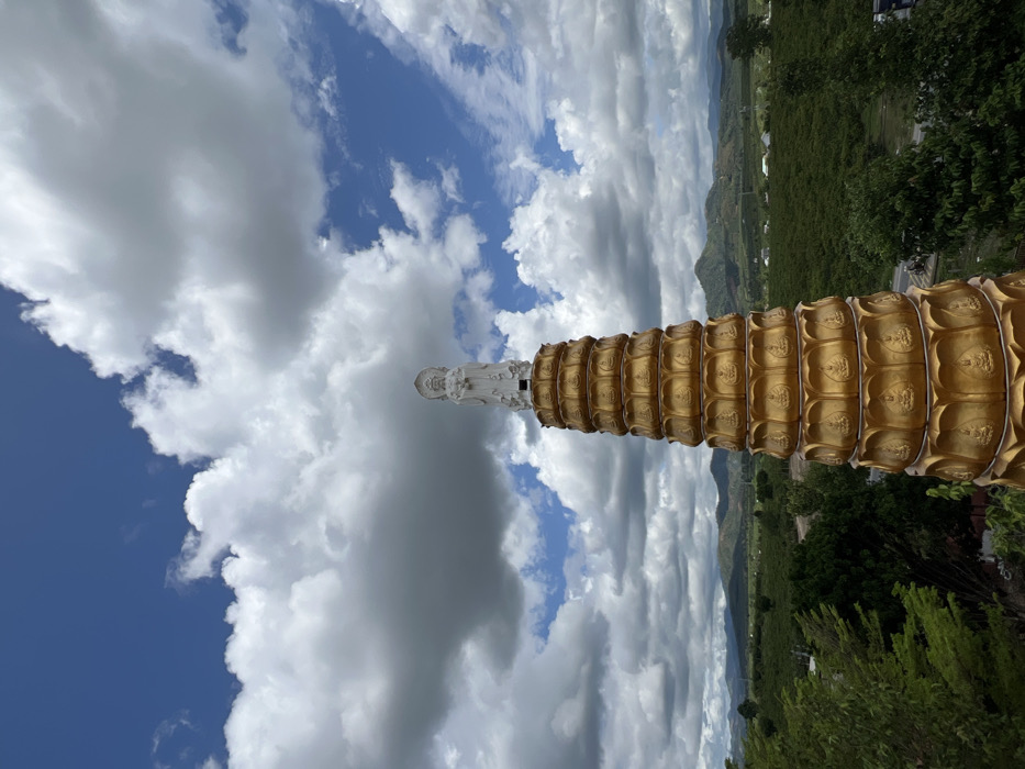 A tall, multi-tiered golden pagoda stands prominently in the foreground, topped with a white statue of a figure resembling Guan Yin, depicted with a halo and holding an object. The pagoda features multiple levels, each adorned with a lotus petal design and small, recessed Buddha figures. The scene is set against a backdrop of rolling green hills and mountains under a partly cloudy sky with patches of bright blue visible through large cumulus clouds. Lush green vegetation, including trees and what appears to be agricultural fields, covers the landscape. A road and scattered structures are discernible in the middle distance. This location is identified as the city of Đăk Mar, Vietnam.