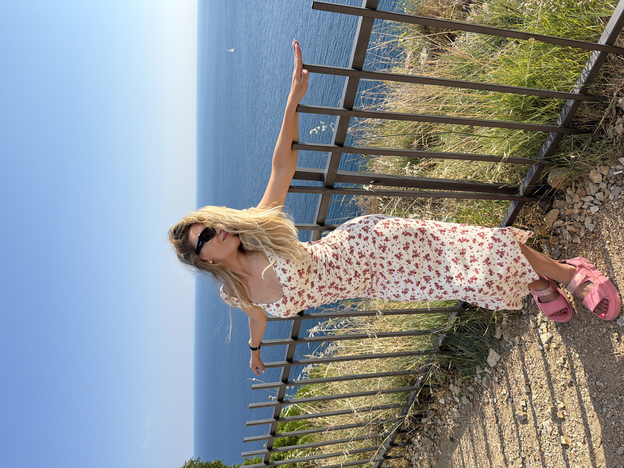A woman with blonde hair and sunglasses leans against a metal fence overlooking the Adriatic Sea. She is wearing a white floral print dress and pink sandals. The background shows a clear blue sky and the blue sea with a small sailboat in the distance. The scene appears to be a scenic overlook, likely in Prijevor, Montenegro, given the vegetation and coastal view. The woman's pose suggests she is enjoying the view and the warm weather. The lighting indicates it is daytime.