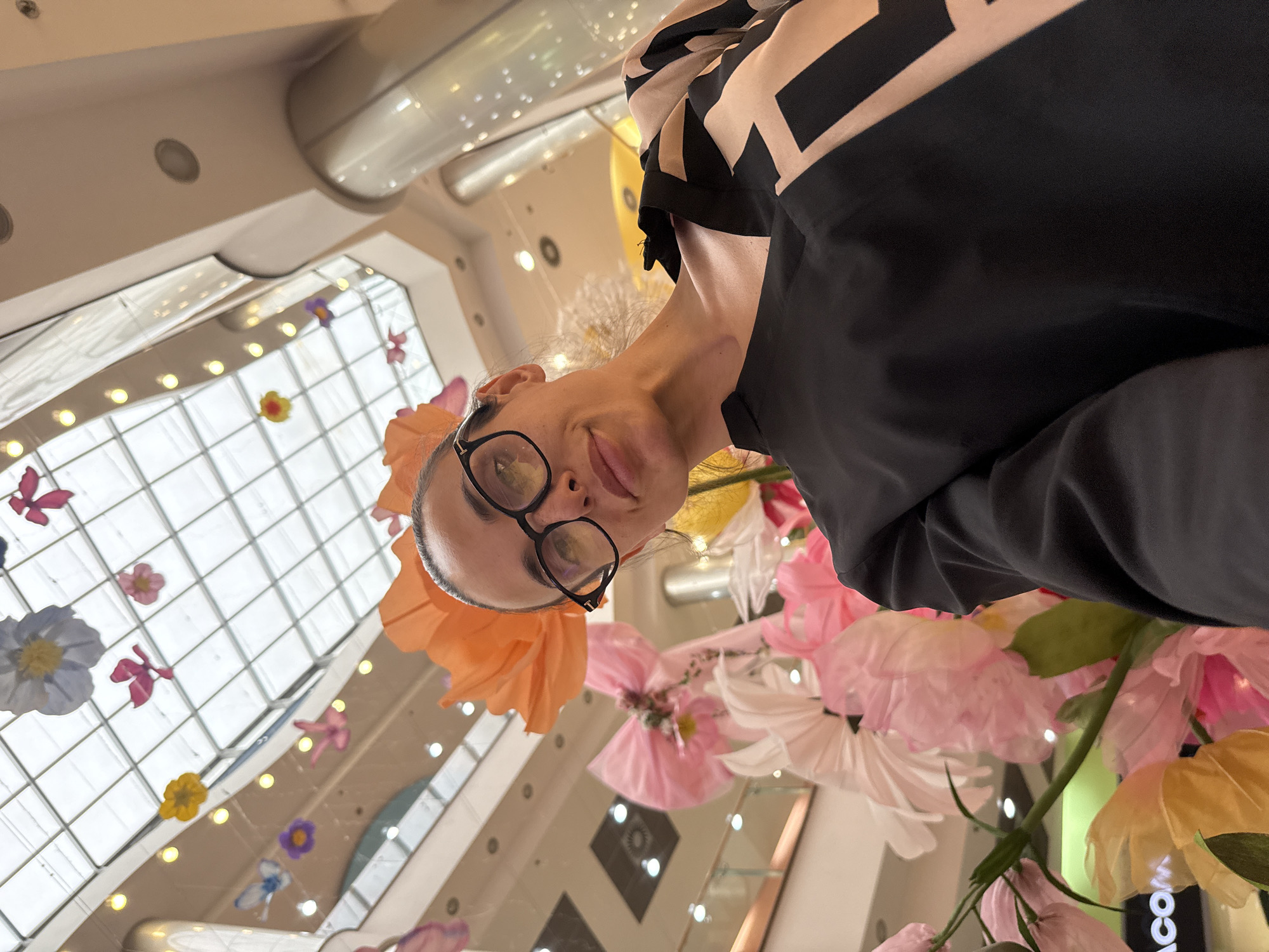 An image taken from a low-angle perspective captures a person, likely a woman, looking directly at the camera. The image is rotated approximately 90 degrees clockwise from its natural upright orientation. She wears black-rimmed glasses, which reflect light, and a black top with a prominent beige and black geometric pattern visible on her left shoulder. Her expression is neutral with a subtle hint of a smile.
The background depicts a brightly lit indoor space, characteristic of a modern shopping mall or large atrium in Pleven, Bulgaria. The setting features a high ceiling with extensive glass panels forming a skylight, allowing ample natural light to illuminate the interior. Numerous circular recessed lights are visible on the white ceiling structures.
Suspended throughout the space are large, colorful decorative elements, primarily paper or fabric flowers and butterflies, in shades of pink, orange, yellow, purple, white, and blue. These decorations create a vibrant, whimsical display, possibly an art installation or seasonal theme. In the lower right corner of the image, a partial view of a dark sign with the text "ACOM" is visible. The person appears to be taking a selfie or being photographed from below, while looking up at the decorative installation.