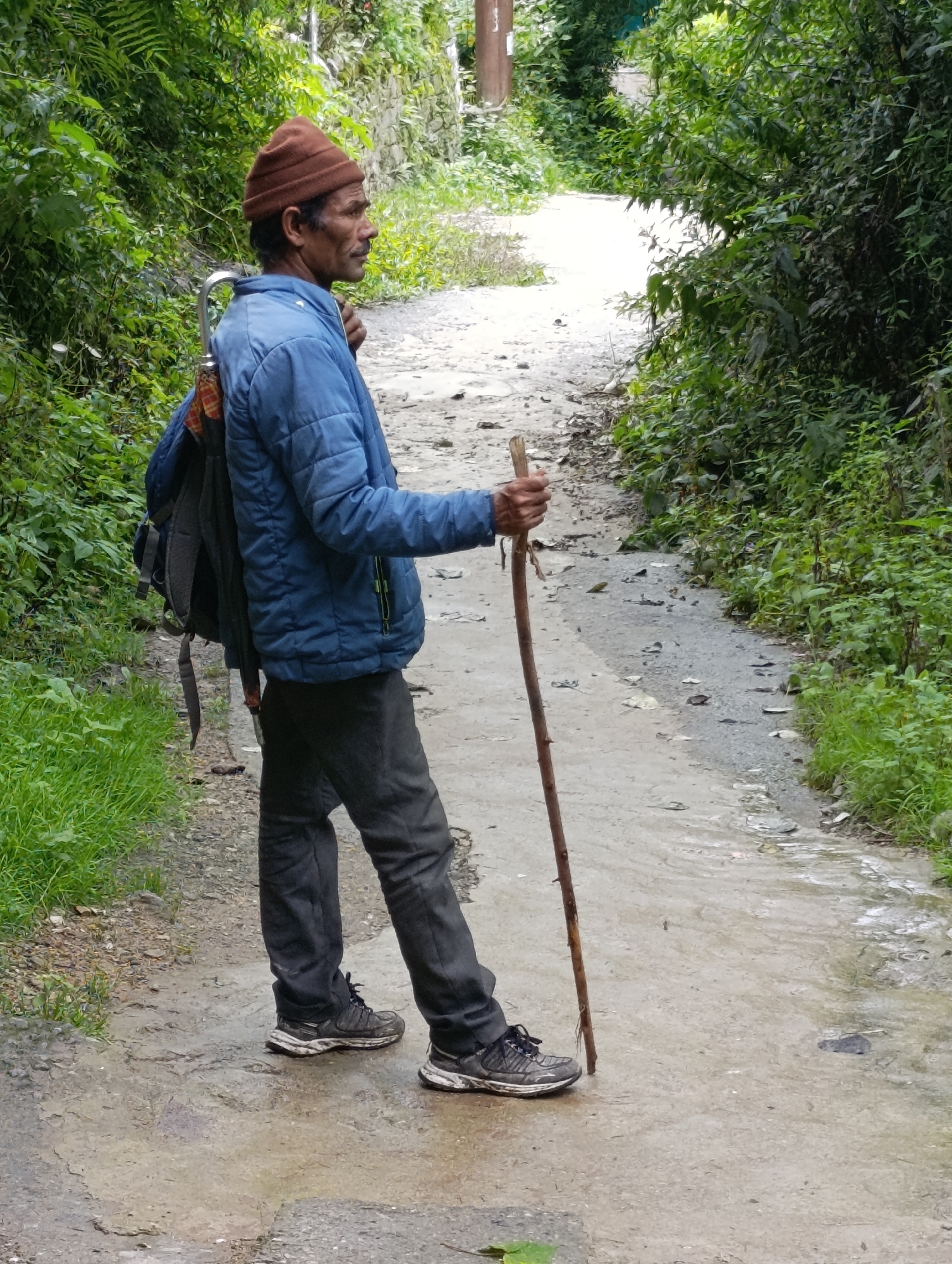 A man in a blue jacket and a brown knit cap stands on a muddy path, holding a walking stick. He wears dark pants and athletic shoes. A backpack is slung over his shoulders. Lush green vegetation borders the path. The path appears to be an unpaved road, possibly in a rural or mountainous area, with water running down it. The lighting suggests an overcast or dim day.