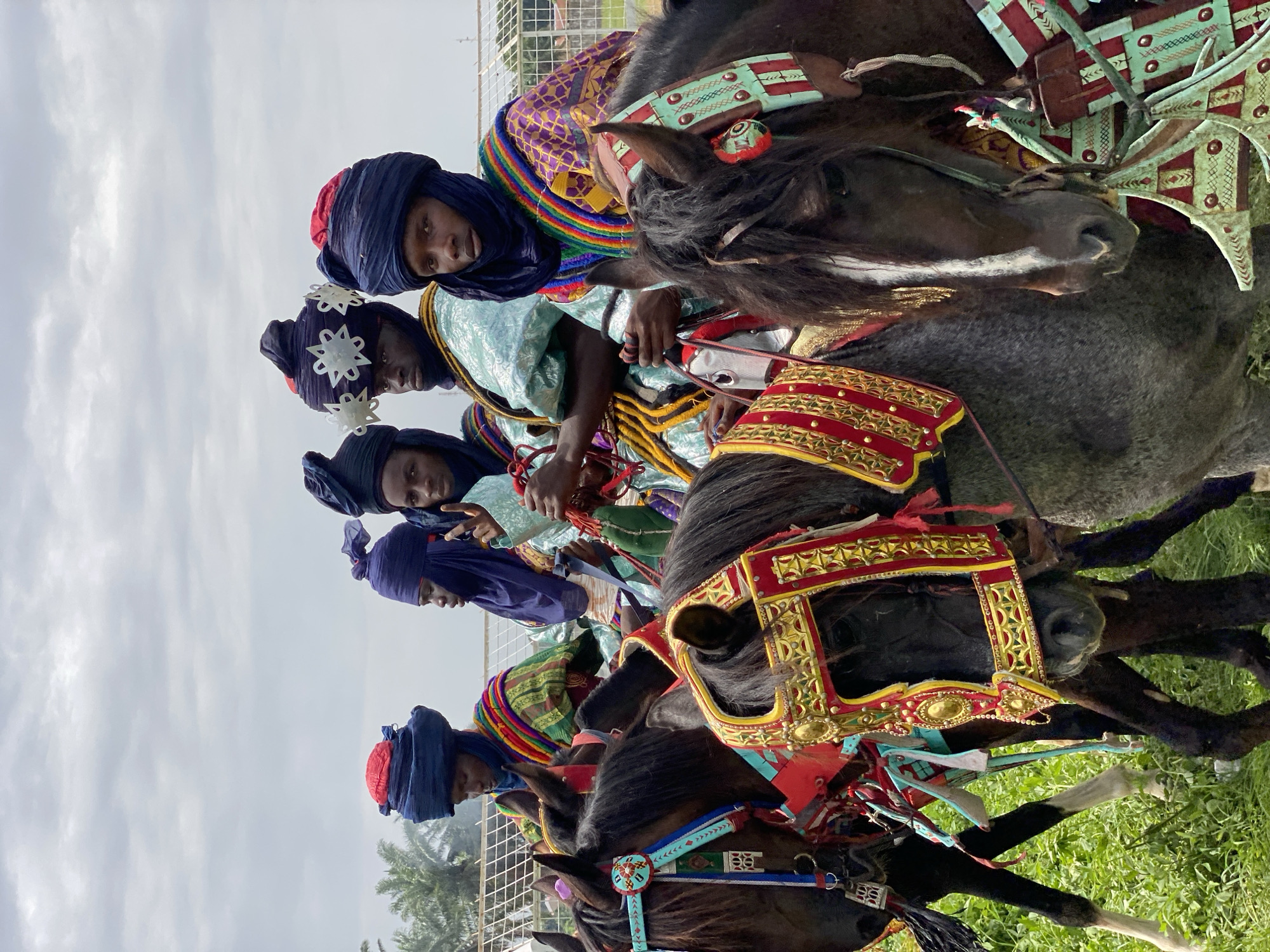 Five men wearing blue turbans and colorful, patterned robes are riding horses that are adorned with ornate saddles and decorative coverings. The men are lined up in a row, facing the same direction. The horses are mostly black with some grey. They are on a green grassy field. The sky is overcast and grey. In the background are some trees and a fence. The scene is a Durbar festival in Kafanchan A, Nigeria.