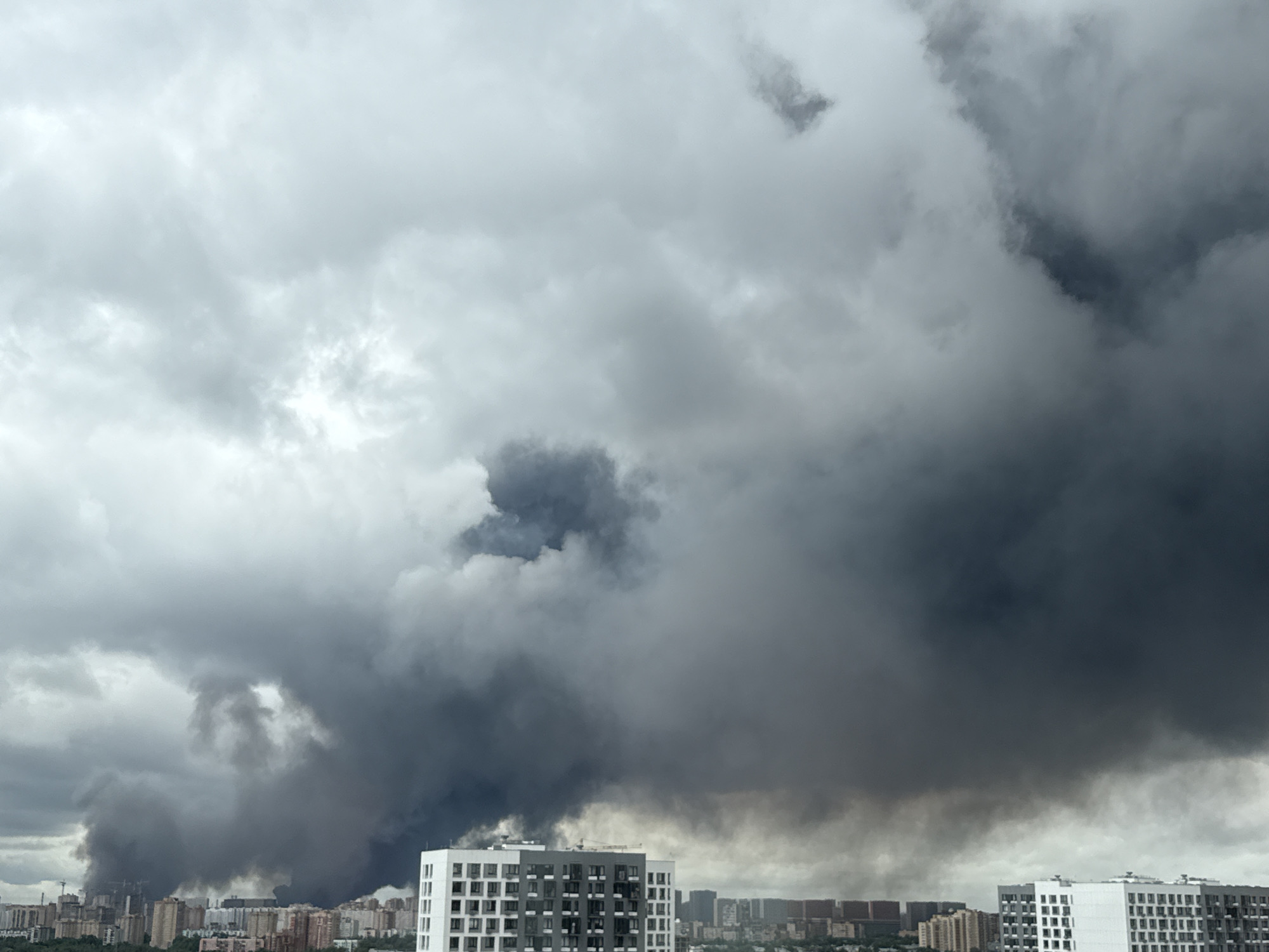 A wide shot captures a panoramic view of the Tomilino, Russia skyline under a heavily clouded, dark sky. The lower portion of the image shows a dense city landscape with numerous residential buildings of varying heights and architectural styles, interspersed with greenery. In the foreground, a modern, white apartment building with numerous windows stands out. Dominating the upper and central portions of the frame is a massive plume of dark, billowing smoke, originating from somewhere within the city and rising high into the atmosphere. The smoke appears thick and ominous, obscuring a large section of the sky and casting a somber mood over the scene. There are no visible people or specific actions depicted, other than the environmental event of the smoke.