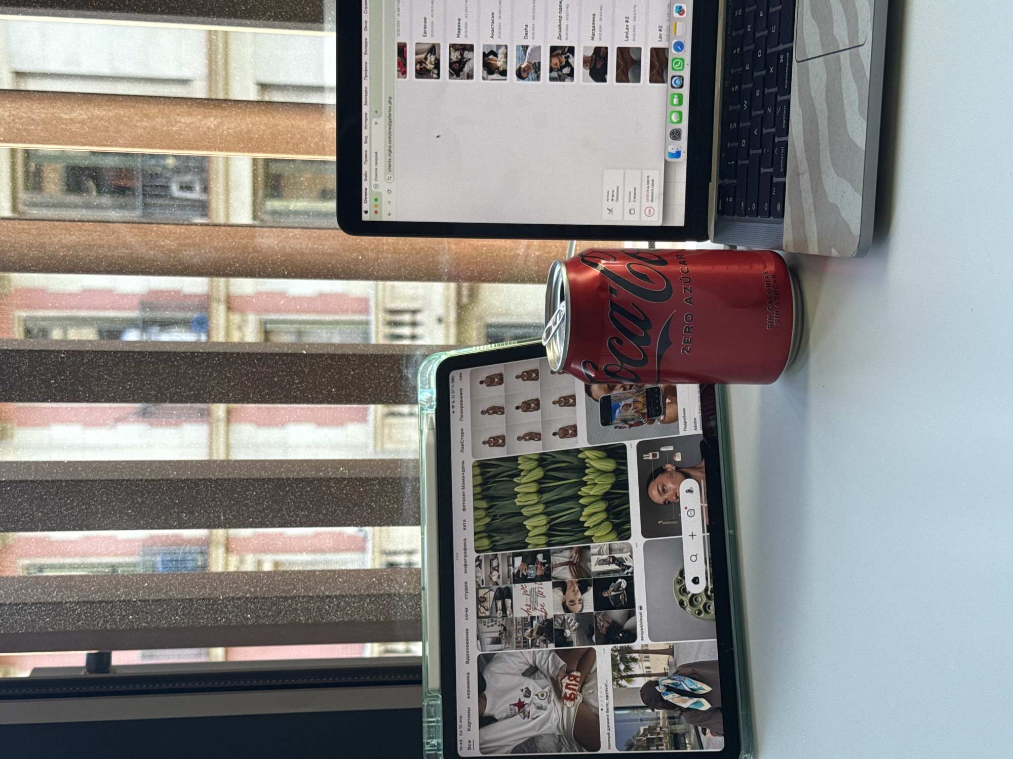 The scene is an indoor workspace. A laptop and a tablet rest on a white desk. The laptop has a zebra-patterned skin. A red can of Coca-Cola Zero sits between the two devices. The laptop screen displays what appears to be a photo gallery interface in a web browser. The tablet displays a Pinterest-style interface with various images. The background features a window with closed blinds, revealing a view of buildings across the street, consistent with the architectural style of Barcelona, Spain. The light suggests it's daytime.