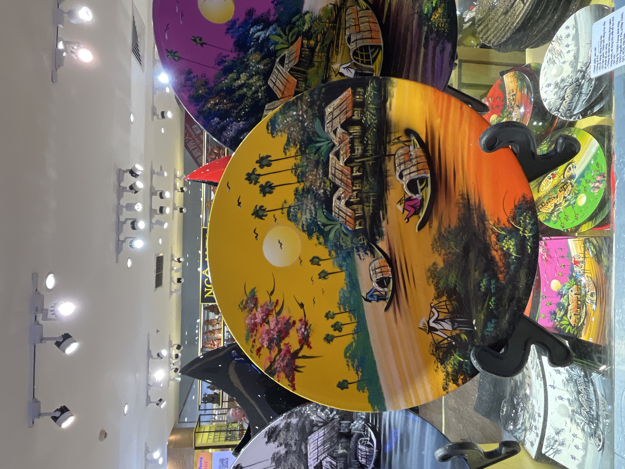 The image shows a display of painted, circular plates, likely tourist souvenirs, possibly located in Nha Trang Ward, Vietnam. The plates depict various landscape scenes, including boats and houses, and are mounted on black stands. The background includes the ceiling with many ceiling lights, shop displays, and other merchandise. The plates feature different color schemes, including yellow, purple, and black and white.