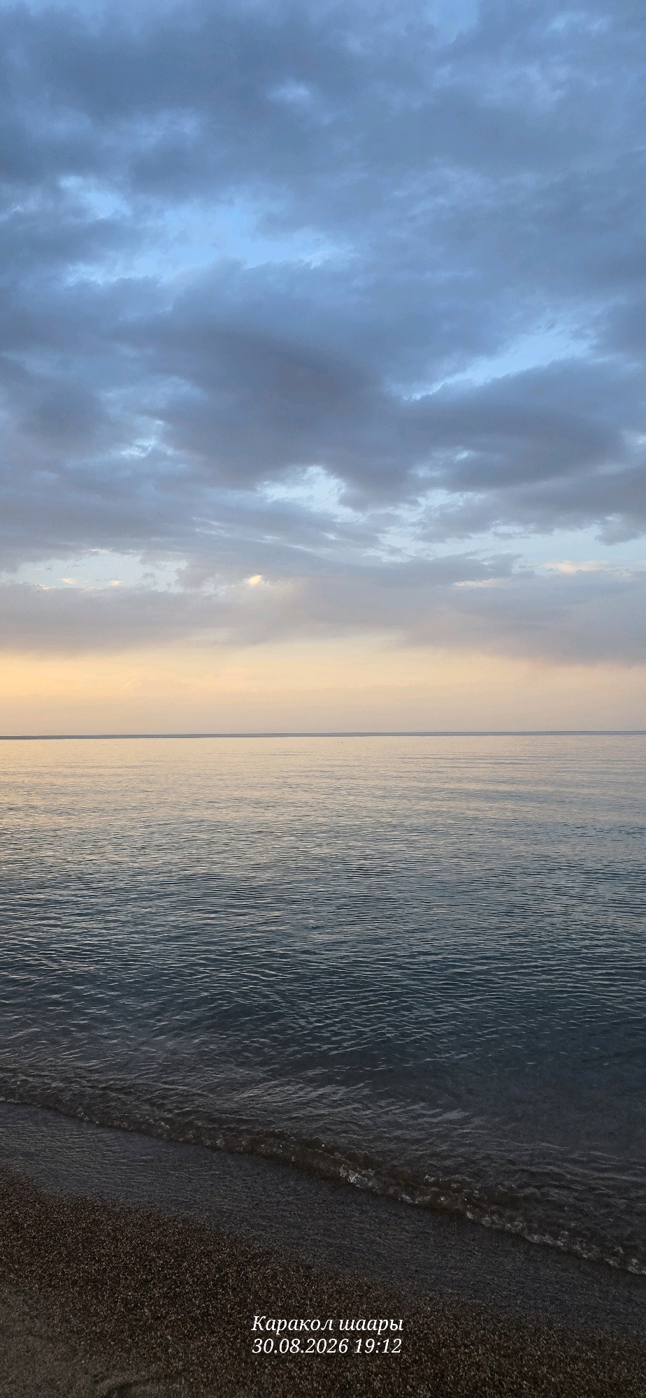 The image depicts a serene seascape at dusk. The sky is a canvas of blues and grays, with scattered clouds tinged with the soft hues of sunset, transitioning to a pale yellow glow near the horizon. The sea is calm, with gentle ripples reflecting the twilight sky. The foreground shows a dark, pebbly shore where small waves lap at the edge of the water. In the lower right corner, text reads "Каракол шаары 30.08.2026 19:12", indicating the location as Karakol and the date and time as August 30, 2026, at 19:12. The scene is tranquil and devoid of human presence, capturing a moment of natural beauty at the end of the day.