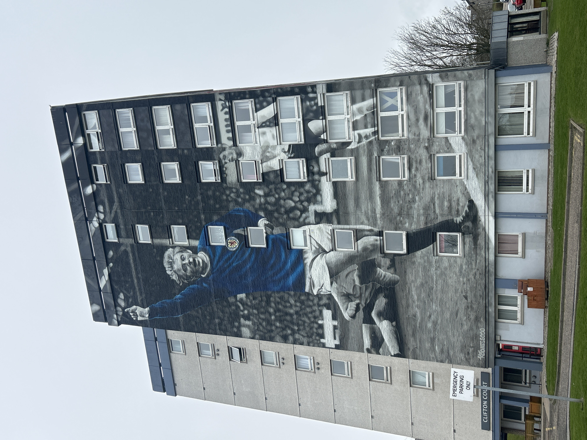 The image shows a high-rise building in Aberdeen City, United Kingdom, featuring a mural of a football player on its exterior. The mural depicts the player in a blue jersey with a team emblem, seemingly in the middle of a game. The mural is predominantly grayscale with the player's jersey highlighted in blue. The background shows a blurred stadium or playing field. The building has multiple rows of windows, many of which appear to be directly incorporated into the mural design. A sign reading "EMERGENCY PARKING ONLY" and "CLIFTON COURT" is visible in the foreground. A patch of green grass is to the right of the building and the sky is overcast.