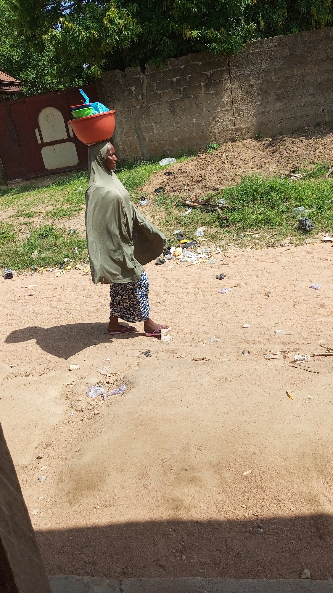 A woman walks along a dirt path, carrying a large orange bowl filled with items on her head. She is wearing a green headscarf and a patterned skirt, and pink flip-flops. The scene appears to be an outdoor market or roadside in Jalingo, Nigeria. The path is sandy and littered with trash, including plastic bags and wrappers. In the background, there is a brick wall and some green foliage. The sun is shining, casting a shadow of the woman on the ground, suggesting it is daytime. The overall impression is one of daily life and commerce in a rural or semi-urban environment.