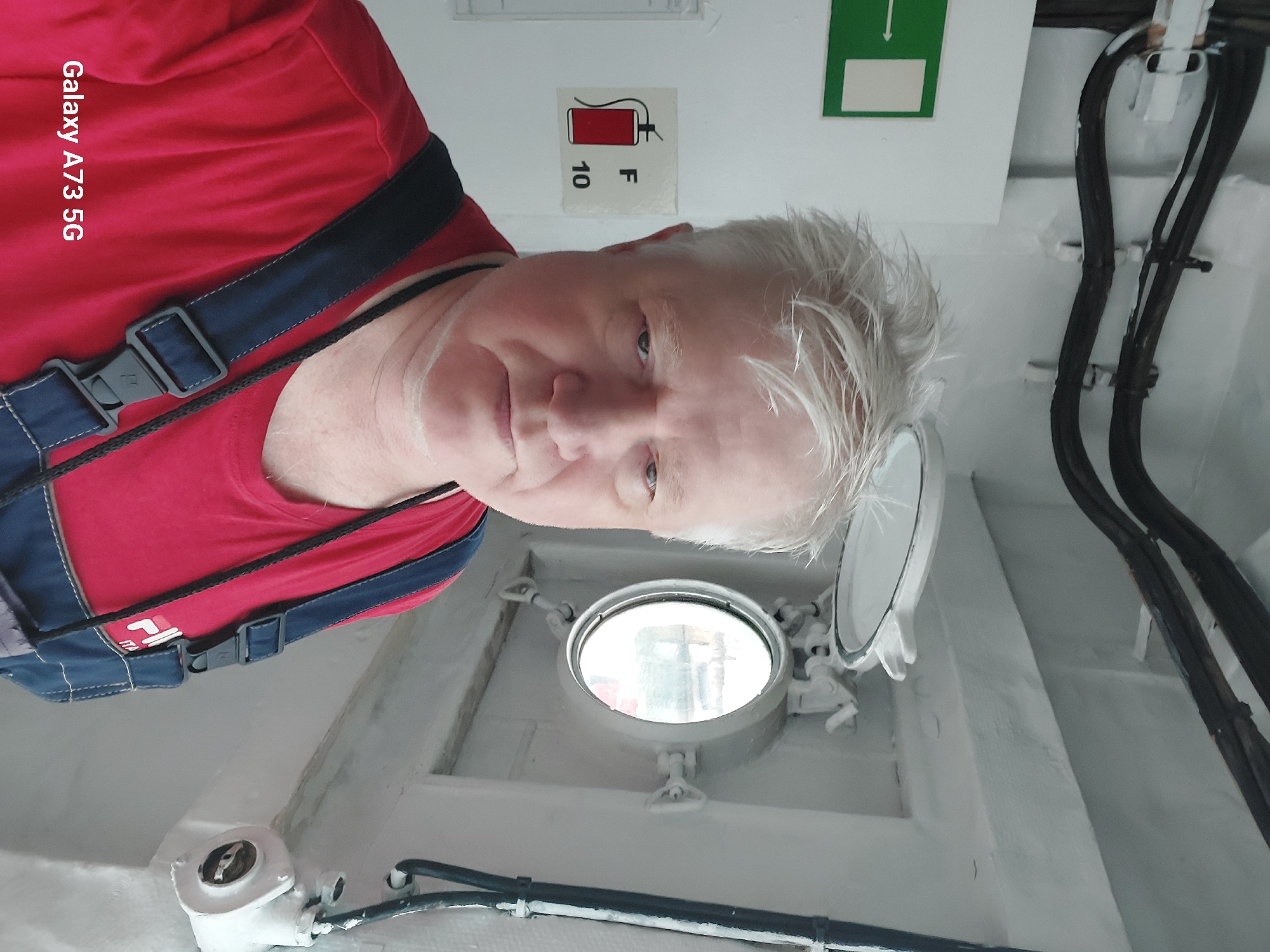 A man with white hair is taking a selfie on what appears to be a ship. He is wearing a red shirt and blue overalls. Behind him, there is a porthole window, some pipes on the ceiling, and a fire extinguisher sign on the wall. The text "Galaxy A73 5G" is in the bottom left corner of the image, indicating the device used to take the photo.
