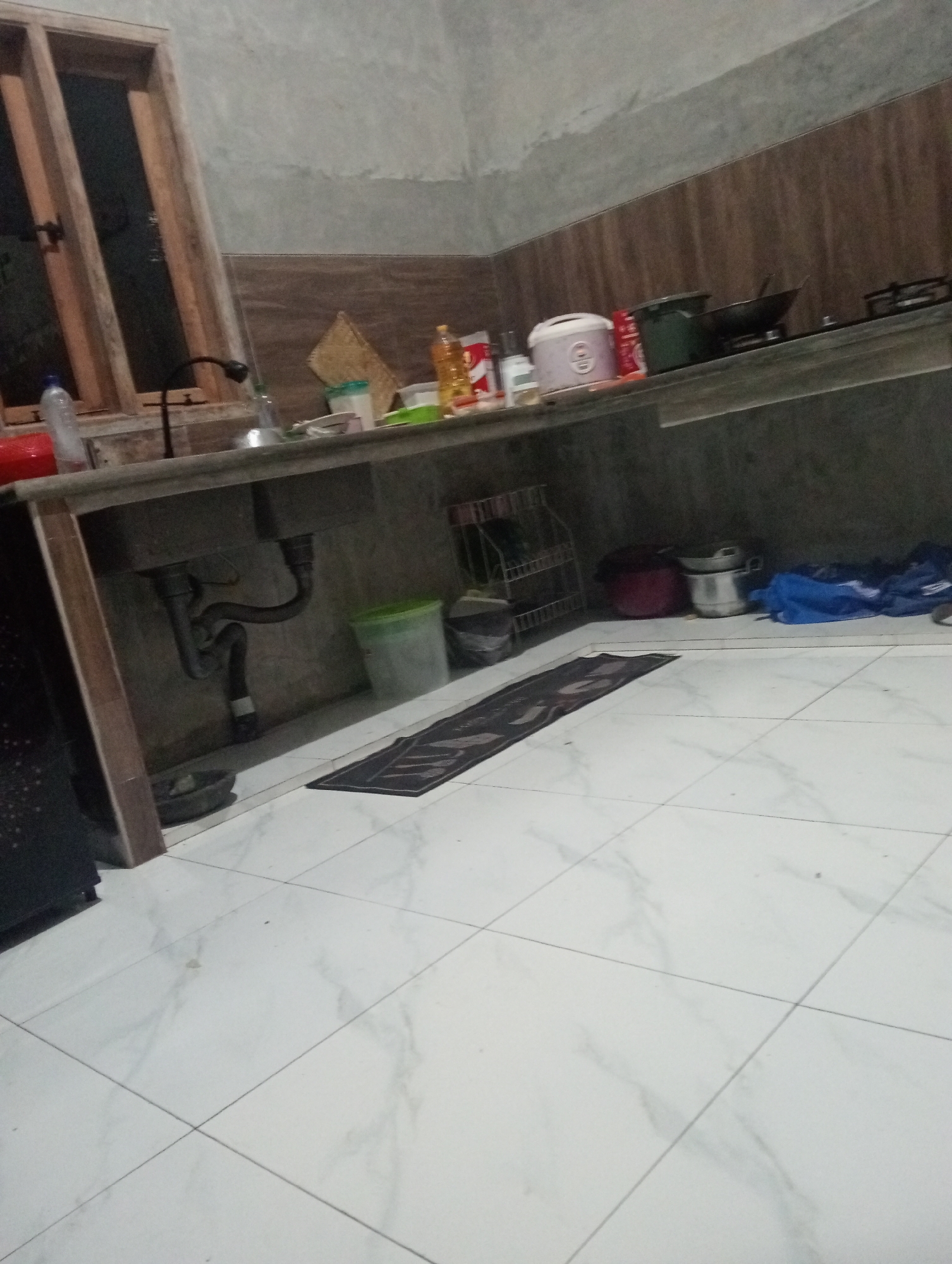 The image depicts a kitchen scene in Muara, Indonesia. The kitchen features a concrete countertop with a sink and a faucet. Various kitchen items are present, including bottles of cooking oil, a rice cooker, pots, and pans. A gas stove is visible on the countertop. The flooring is tiled, and a dark rug is placed on the floor. The walls are plastered, and there is a wooden window frame. The scene appears to be dimly lit, suggesting it might be evening or a poorly lit indoor space. There are no people visible in the image.
