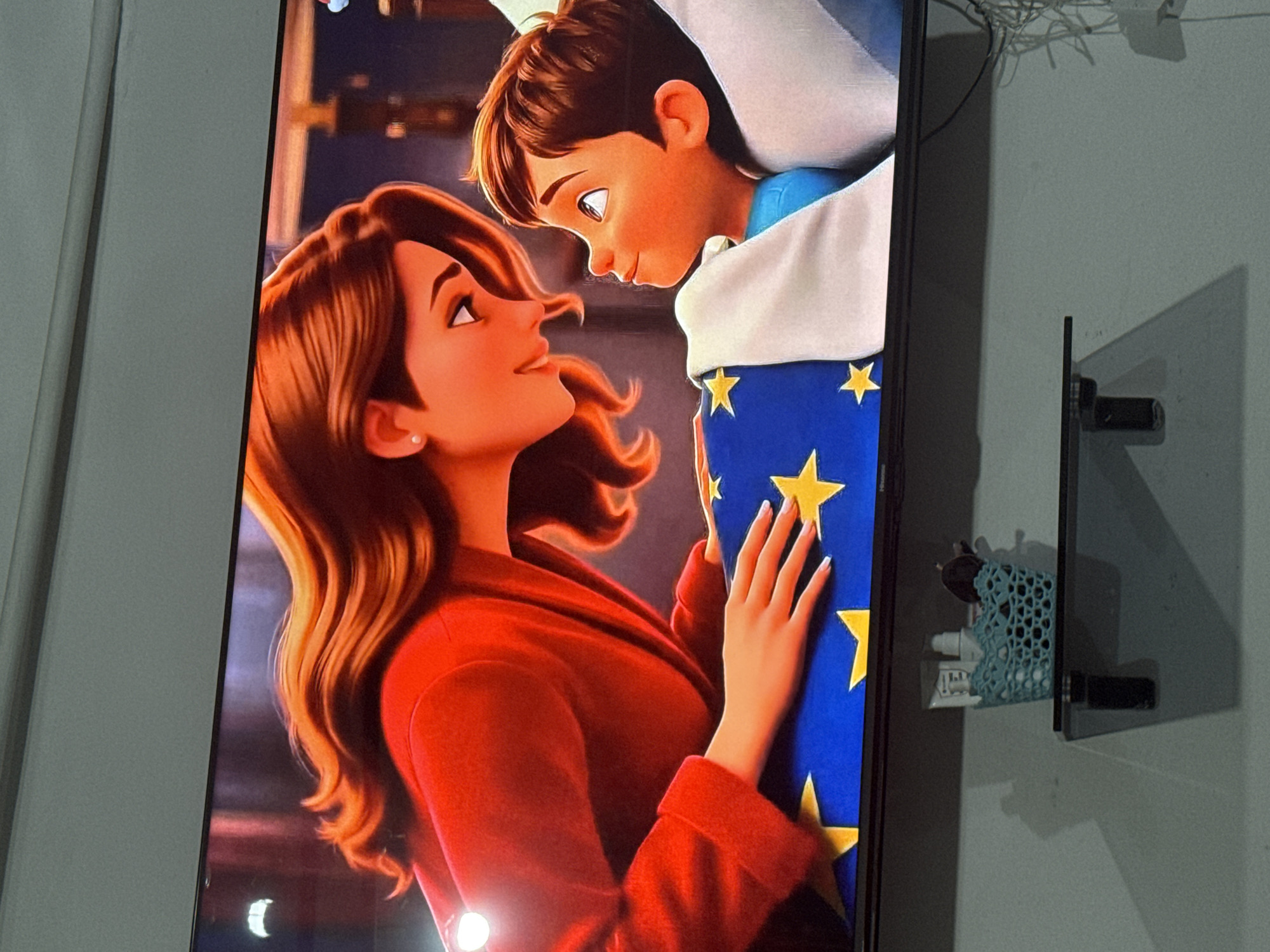 The media file shows a close-up of an animated scene from a screen, possibly a television or computer monitor, against a neutral gray wall. The scene depicts two animated characters: a woman with long, wavy brown hair and a red jacket, and a young boy with brown hair. They are looking at each other with affectionate expressions. The woman is reaching her hand towards the boy, who appears to be nestled in a blue blanket adorned with yellow stars. The lighting in the scene is warm, suggesting an indoor setting, possibly a bedroom. The image is captured from a slight angle, and to the right of the screen, a portion of the wall is visible, with a black mount and a decorative blue object casting shadows. The overall mood of the animated scene is tender and intimate. There is no visible text in the media file.