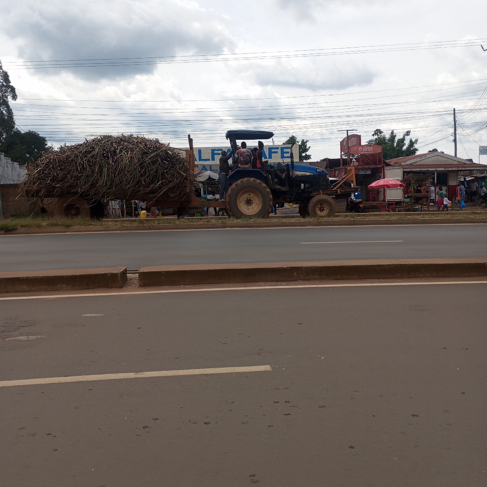 The image depicts a blue New Holland 1002 tractor towing a trailer loaded with what appears to be a large amount of dried plant material, possibly sugarcane, on a paved road. Several individuals are riding on the tractor. The scene is in Sheywe ward, Kenya, as evidenced by the background buildings, including establishments such as "ALPHA CAFE" and a cellular service retailer "Airtel" alongside "M-PESA," and a butchery. People are visible near the buildings, suggesting commercial activity. The sky is overcast. The road surface is brown and exhibits some wear and tear, with a white line dividing lanes. A low concrete barrier separates the road from the immediate roadside.