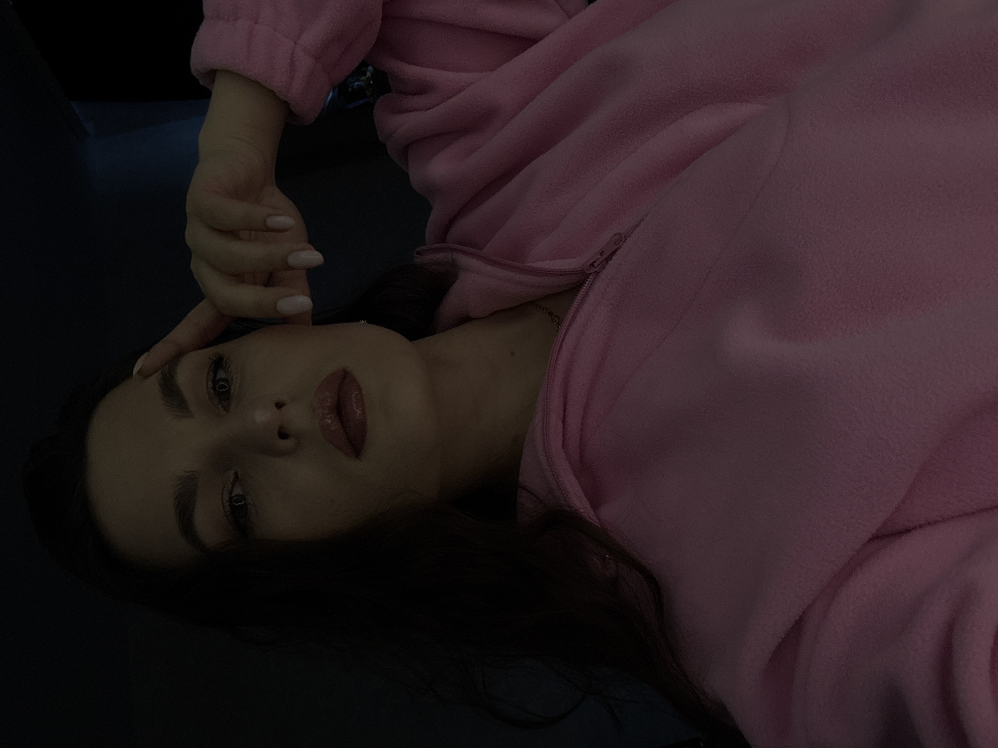 A woman is pictured in a close-up, self-taken photograph, likely indoors, given the dim lighting. She has dark hair and is wearing a pink zip-up fleece jacket. Her right hand is raised to her forehead, with her index finger touching her temple. Her makeup is visible, featuring prominent lipstick. The background is dark and indistinct. The location of Chaikovsky, Russia is not ascertainable from the image.