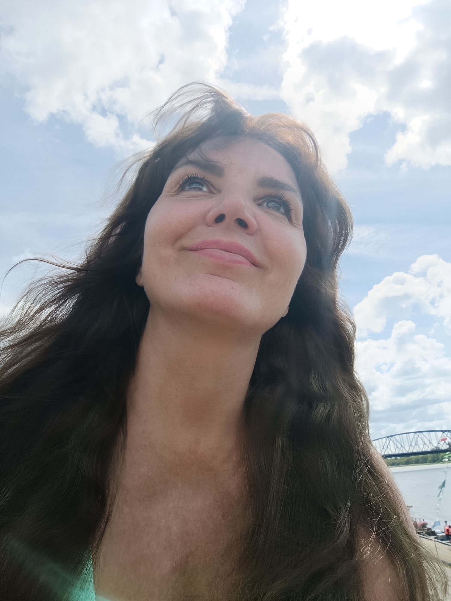 This is a low-angle, waist-up, medium shot of a woman outdoors with a bright, cloudy sky behind her. The woman has long, dark brown wavy hair that blows gently in the wind. She has fair skin with light makeup and her head is tilted upwards and to the right, with her gaze directed towards the sky. She is smiling slightly, with her lips slightly parted. The background features a body of water, a bridge in the distance, and some white structures that appear to be tents or sails. There are no identifiable landmarks that place this image specifically in Toruń, Poland. The lighting suggests it is daytime, possibly late morning or afternoon, with the sun casting a soft glow and some lens flare on the left side of the image.