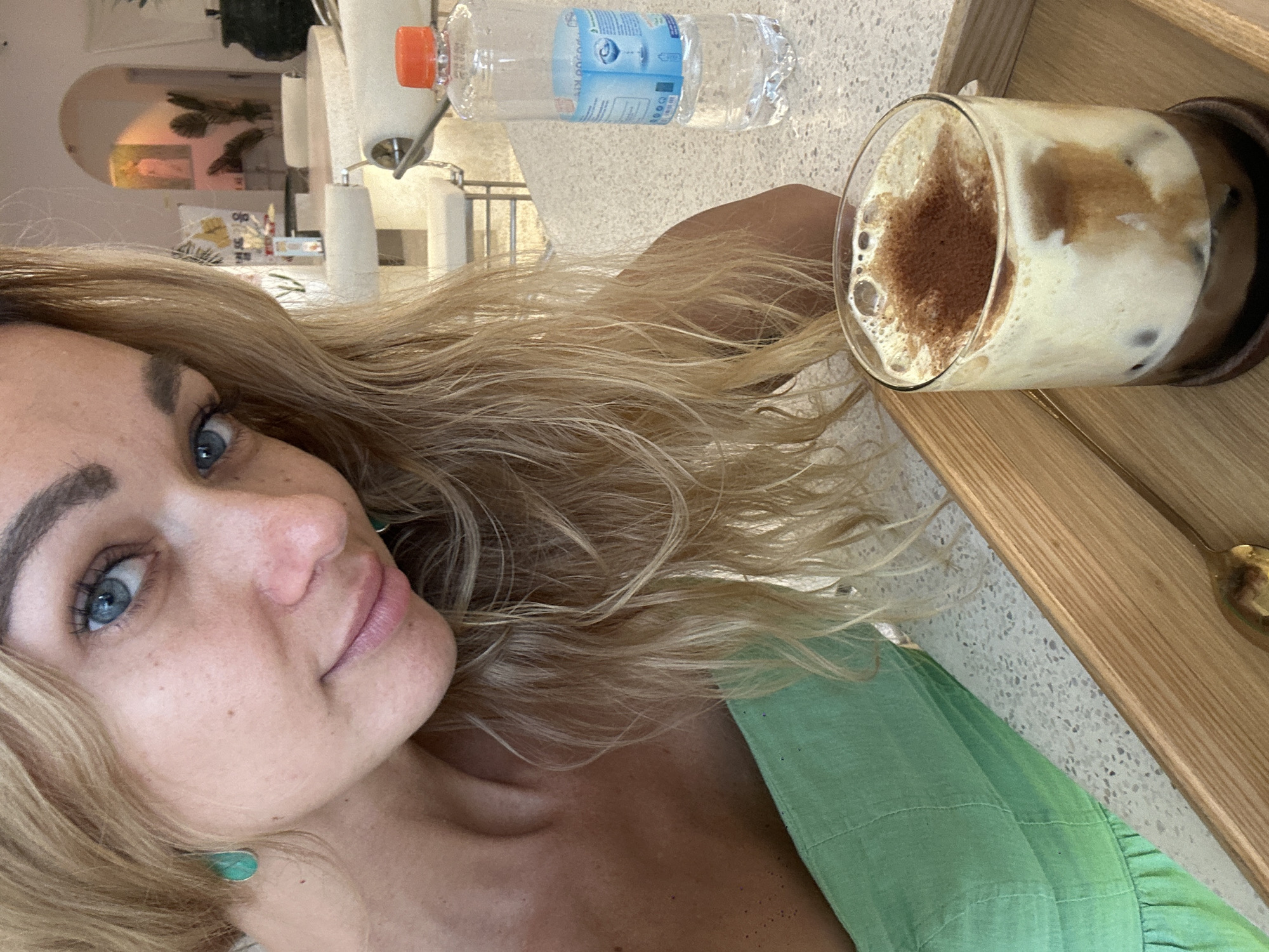A woman with blonde hair and blue eyes is taking a selfie. She is wearing a green shirt and turquoise earrings. A glass of iced coffee with froth and a sprinkle of cocoa powder sits on a wooden tray next to her. A water bottle also sits on the countertop. The background suggests a bright, airy indoor setting. Without further information, the exact location within Phu Thuy Ward, Vietnam cannot be determined.