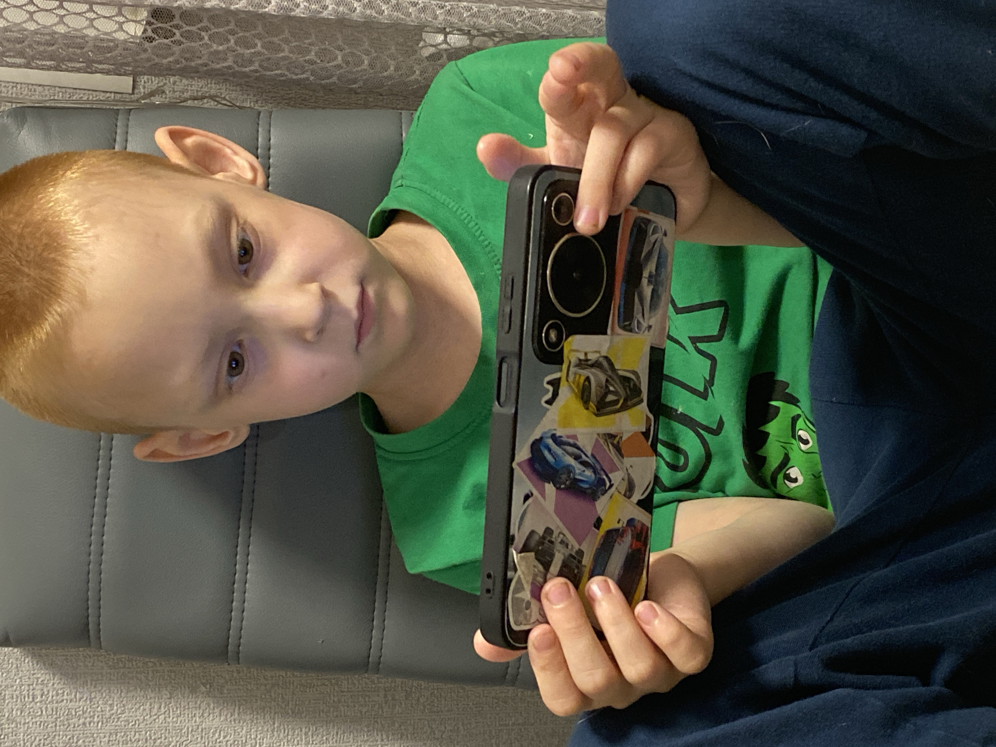 A young boy with fair skin, light brown hair, and brown eyes is depicted in a medium shot, lying down or reclining indoors. He is wearing a bright green t-shirt with a cartoon character, likely the Hulk, visible on the side, and dark blue pants. He is holding a black smartphone with both hands, positioned in front of his chest.
The smartphone is encased in a black protective cover that features a prominent camera module at the top. The back of the phone case is decorated with numerous small, colorful stickers or cutouts, primarily depicting various car designs, including sports cars and race cars. The boy's attention appears to be directed forward, possibly towards the phone screen (which is not visible) or slightly beyond it, with a neutral and engaged expression.
He is resting on a gray, upholstered surface with vertical stitching, resembling a sofa or a headboard. In the background, behind his head, there is a light-colored, patterned fabric, possibly curtains or a textured wall covering, with a lace-like appearance. The lighting suggests an indoor setting, likely with artificial illumination. No specific time of day or weather conditions can be determined from the image. The location is contextualized as Noviy, Russia. No discernible text is visible in the frame.