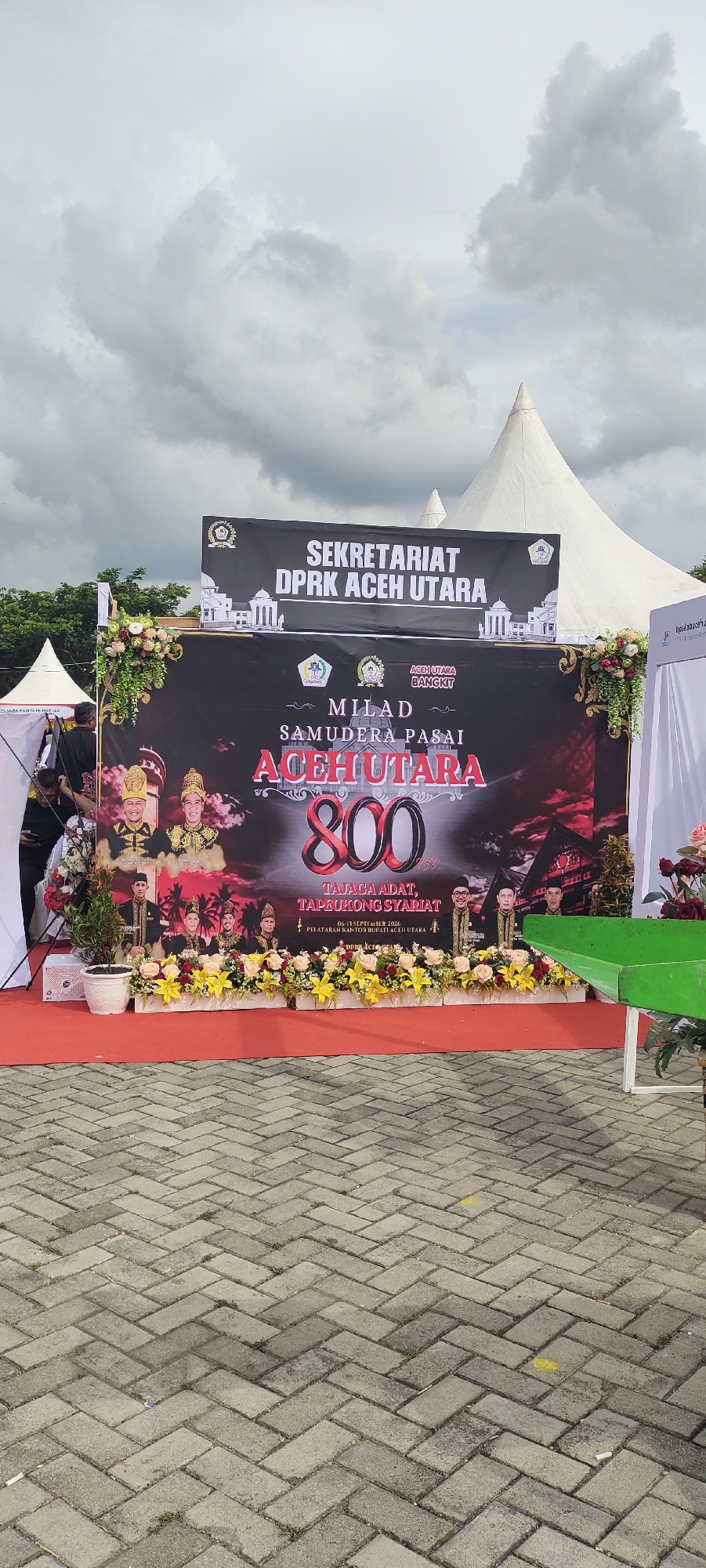 A banner displayed outdoors on a cloudy day features the text "SEKRETARIAT DPRK ACEH UTARA" at the top. Below, it reads "MILAD SAMUDERA PASAI ACEH UTARA 800 TAHUN" and then "TAJAGA ADAT, TAPEUKONG SYARIAT". The banner also includes images of men in traditional attire and floral decorations. In front of the banner, a red carpet is laid over a paved ground, with potted plants and more floral arrangements. To the right of the banner, part of a white tent and a green metal structure are visible. The setting appears to be an outdoor event in Meunasahaluedrienlhoksukon Barat, Indonesia.