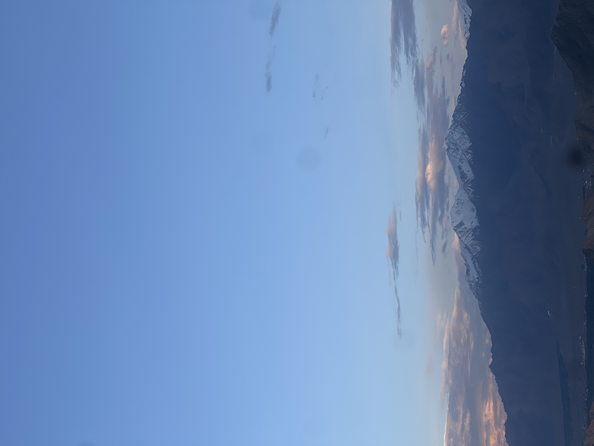 The image depicts a panoramic view of the city of Leh, India, nestled at the foothills of snow-capped mountains under a clear blue sky. The foreground is dominated by the vast expanse of the sky, with a few scattered clouds tinged with the soft hues of dawn or dusk. Below the sky, a range of majestic mountains rises, their peaks dusted with snow. The lower slopes of the mountains transition into the dark silhouette of the Leh valley, where the faint outlines of buildings and possibly a settlement are visible. The overall impression is one of serenity and grandeur, capturing the breathtaking landscape of this high-altitude region.