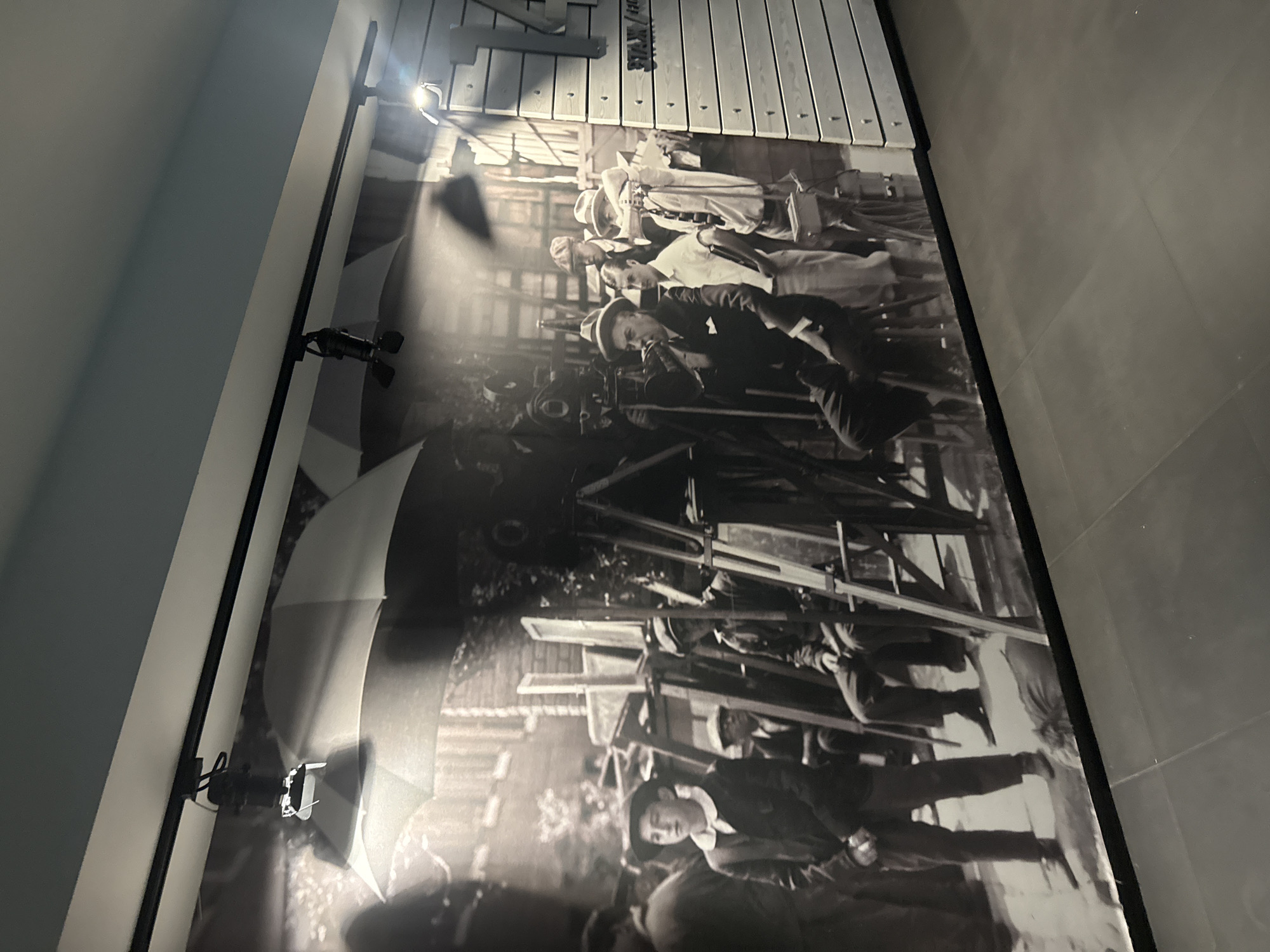 The image is a photograph of a large mural depicting a scene from early filmmaking. The mural features several men in period clothing, some of whom appear to be involved in operating film equipment such as cameras and lights. There are also large, white umbrella-like diffusion screens casting soft light on the scene. The mural is installed on a wall in a modern interior space, indicated by track lighting above the mural and a tiled floor below. On the right side of the mural, a section of the wall appears to be constructed of horizontal wooden planks, with some text visible. The overall impression is of a historical exhibit related to cinema. The location provided, "город Ялта, Ukraine," suggests this mural might be displayed in a museum or public space in Yalta.