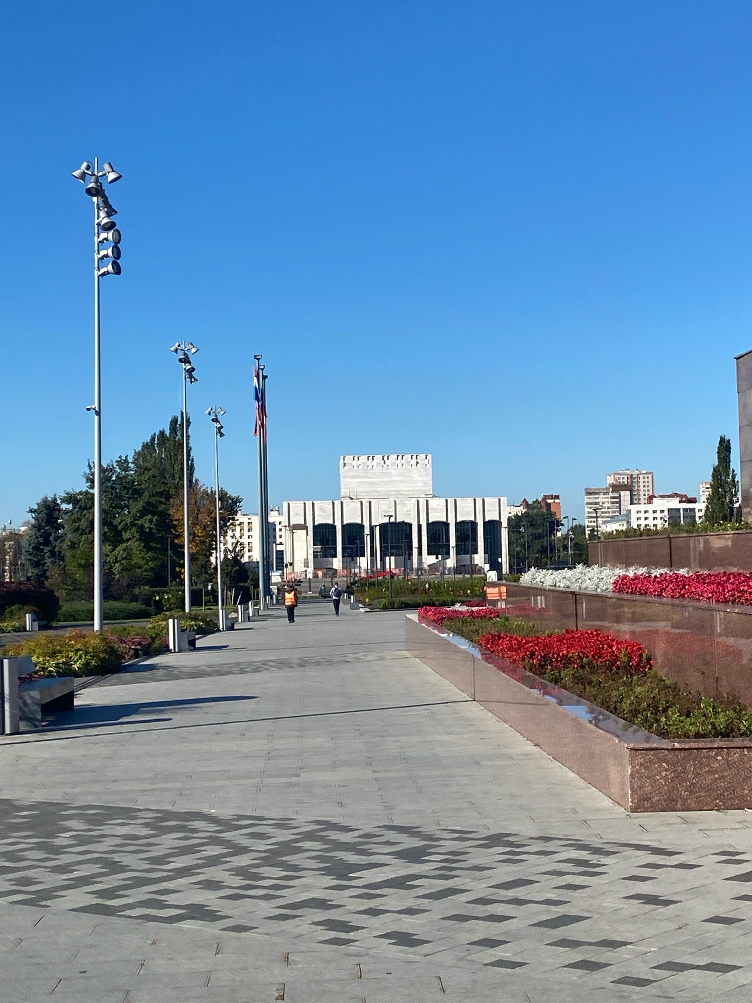 The image depicts a wide, paved pedestrian walkway leading towards a large, modern building with a classical facade. The walkway is lined with planters containing red and white flowers and green foliage, as well as several benches. Tall lampposts with multiple lights are also visible along the walkway. In the background, trees and other city buildings can be seen. Two individuals are walking on the path, one wearing a bright orange vest. The sky is clear and blue. The scene appears to be set in a public square or park area in the city of Perm, Russia, specifically in front of the Perm Academic Theatre.