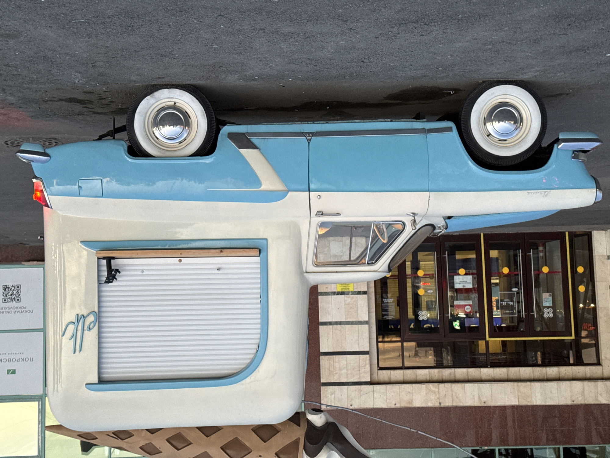 A vintage, two-tone blue and white truck with a food truck conversion is parked on a paved surface. The truck has a rounded, cream-colored rear section with a blue-trimmed window that has a white, rolling metal shutter. The word "elk" is written in cursive blue lettering on the side of the rear section. The truck is parked in front of a building with a brown, textured facade and several glass doors, some of which are marked with yellow circles and signs. The building appears to be a commercial establishment. There are signs on the building, one of which has the text "ПОКРОВСКИЙ" and a green logo. The ground is asphalt, with some dark, wet patches. The lighting suggests it is either early morning or late afternoon. The scene is likely a street in Yekaterinburg, Russia.