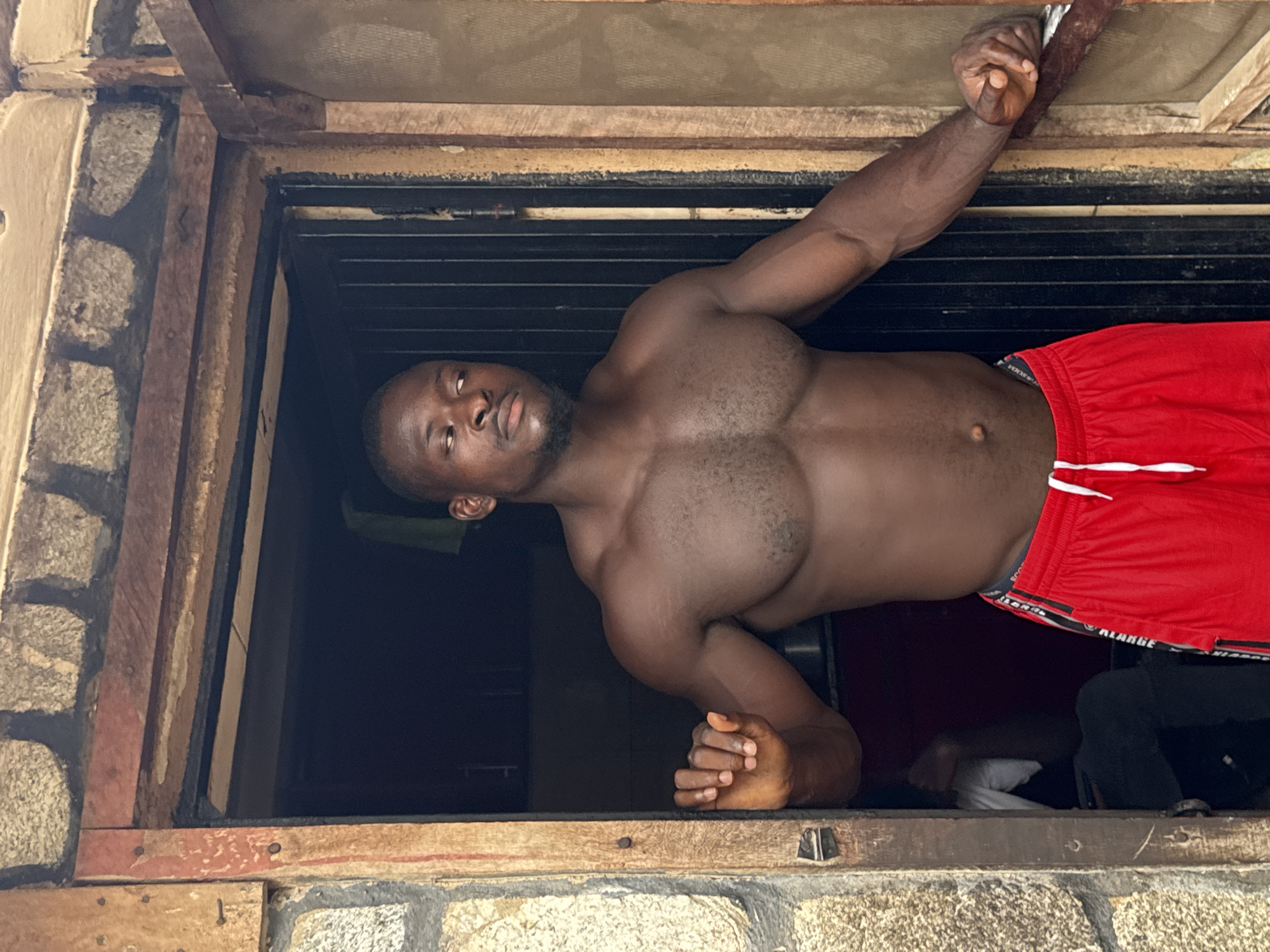 A dark-skinned man with a muscular build stands in a doorway, looking to his right. He is shirtless and wearing red shorts with white drawstrings. The doorway is framed by rough, gray stone and weathered wooden planks. Behind him, a dark interior is visible with a black metal gate or grill. To his left, another person can be seen in the background, sitting down and wearing dark clothing. The setting appears to be a simple structure, possibly a residential building or shop, in Akwanga, Nigeria.
