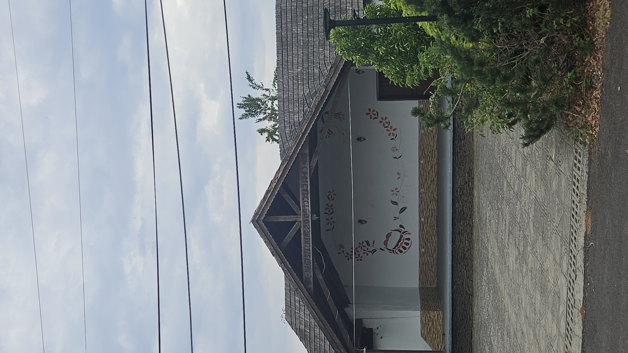 The image depicts the facade of a building with a distinctive roof in Veľké Slemence, Slovakia. The building features a white wall adorned with floral motifs and a basket design. Above the wall, exposed wooden beams are visible under the eaves of a steeply pitched, shingled roof. Carved into one of the wooden beams is text that appears to read "NAGYSZENCSE 2005." The building has a porch or covered area in front, with a textured tiled pavement leading to it. To the right of the building, dense green foliage and trees partially obscure the view. Overhead, power lines are strung across the cloudy sky. The overall scene suggests a rural or semi-rural setting, possibly a community building or a traditional house.