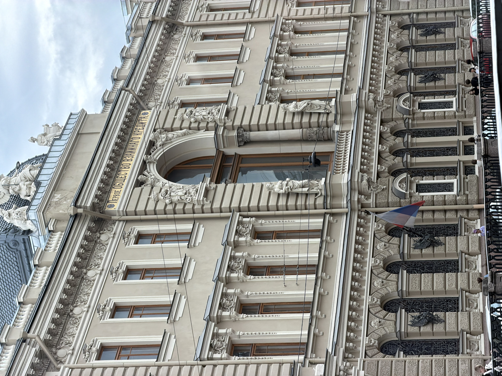 The image shows a detail of a grand, ornate building with classical architectural elements. The facade is a light beige color with extensive white decorative trim. There are multiple stories of windows, many with arched tops. The windows have dark wooden frames. The building features elaborate cornices, friezes with sculpted motifs, and statues or figures adorning the upper levels and around some of the windows. A prominent feature is a large arched window on the upper floor, flanked by columns. Above this window, a decorative inscription in Cyrillic is visible, reading "ПЕРВОЕ ОБЩЕСТВО ВЗАИМНАГО КРЕДИТА." A Russian flag is displayed on a flagpole attached to the building on the right side of the image. The lower portion of the image shows a sidewalk or promenade with a black railing, and several people are visible standing and walking along it. Cars and more railing are visible in the far distance. The sky is overcast. The architectural style suggests a historical public building. The inscription indicates this is the "First Mutual Credit Society." This building is located in Saint Petersburg, Russia, not Lednevo.