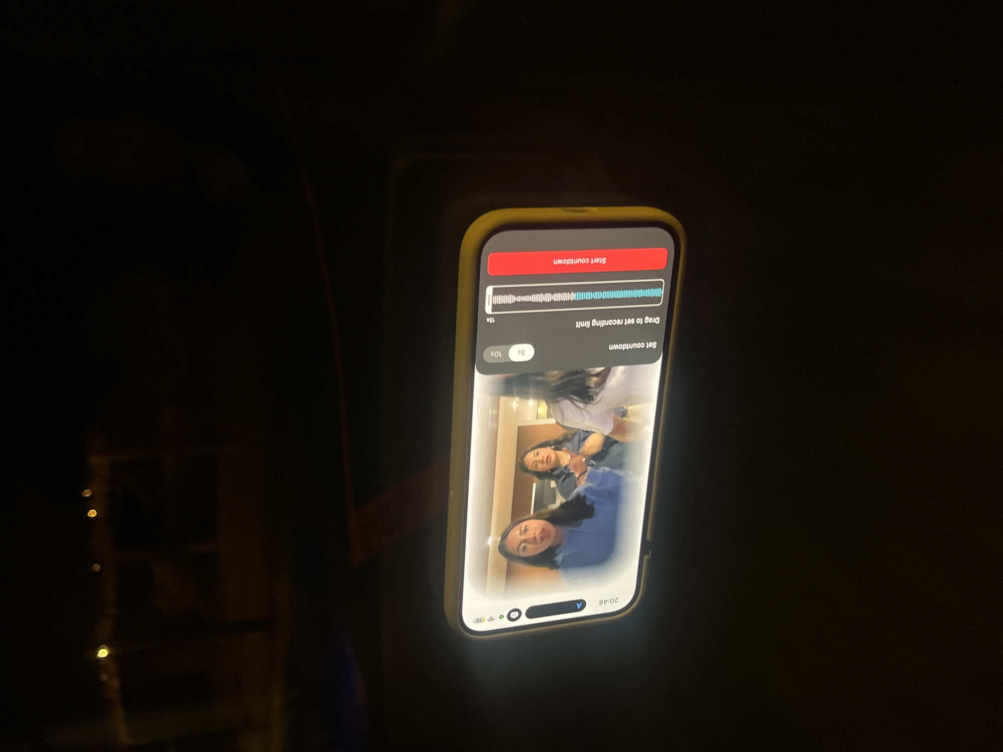 A smartphone with a yellow case is held in a dark indoor environment. The phone's screen displays a video of three people. The person in the foreground is wearing a blue shirt. The phone's screen also shows controls for setting a recording countdown. The time displayed on the phone is 20:48. The location, El Oasis Valsequillo, Mexico, cannot be determined from the image.