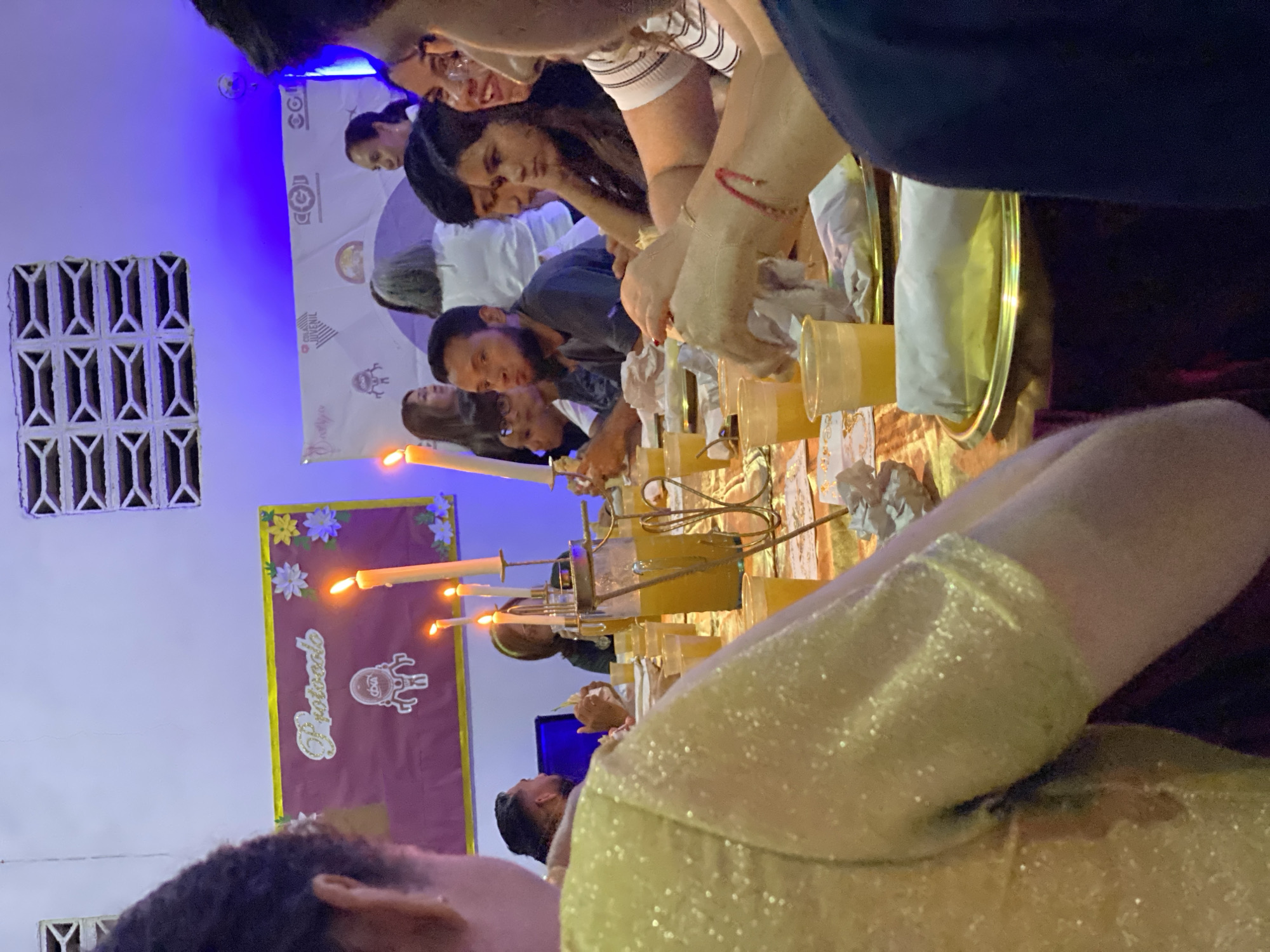A group of people is gathered around a long table covered with a patterned cloth. There are illuminated candles on the table, with what appears to be several clear cups placed along the length of the table as well. The people are looking down at the table, with some holding items that appear to be paper or cloth and placing them into the cups. The scene appears to be taking place indoors. A banner on the wall with floral decorations has the word "Protocolo" written on it. A second banner behind the table includes a logo that contains the text "Juvenil". The location is Tinaquillo, Venezuela. A window with a distinctive design can be seen to the left of the banners.