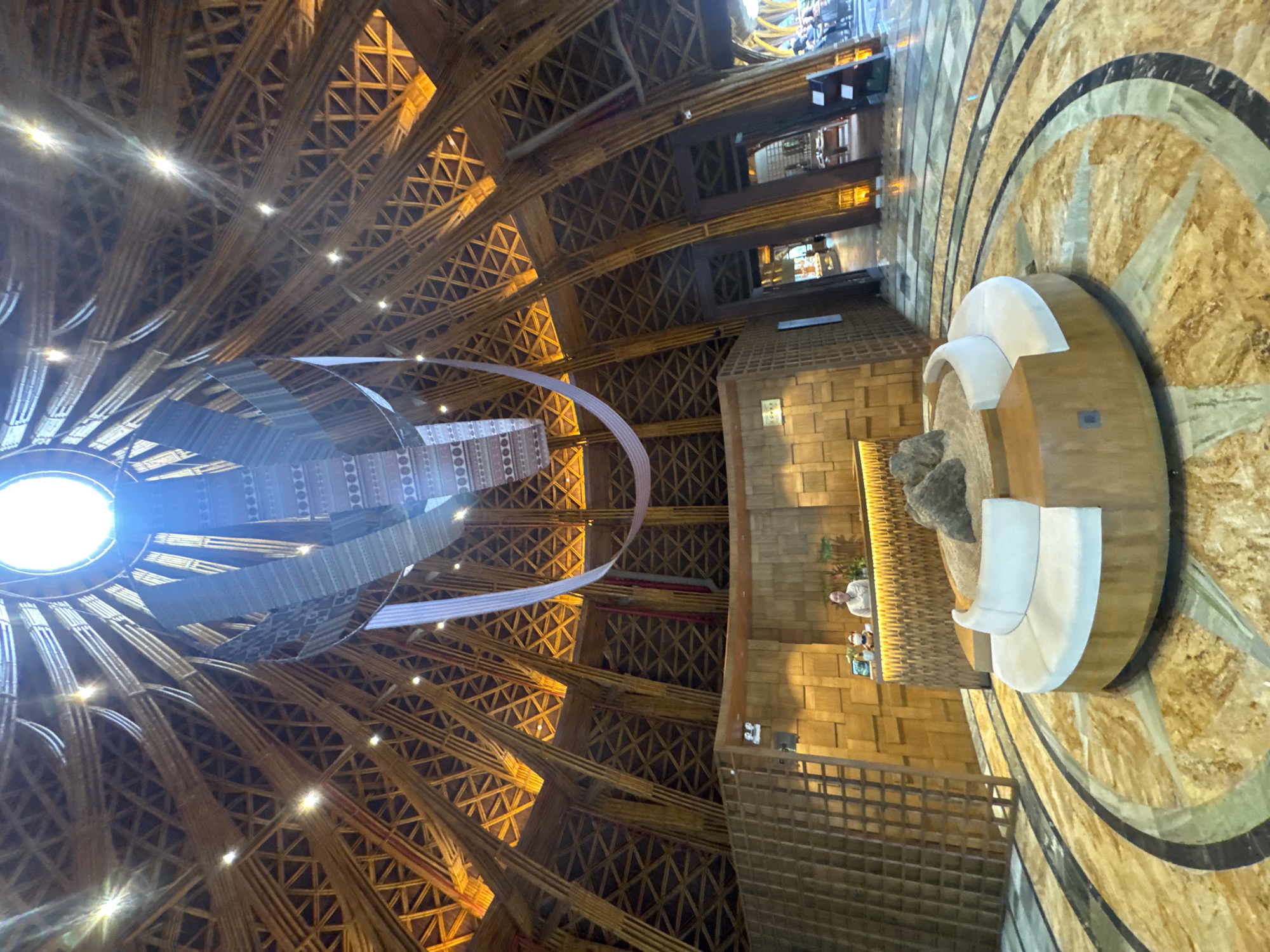 This image captures the interior of a large, high-ceilinged building in Púng Luông, Vietnam, consistent with a resort lobby or public space. The dominant feature is the vaulted, conical ceiling constructed from numerous light-colored wooden or bamboo beams, forming a complex lattice pattern of interconnected triangles and diagonals. Multiple warm-toned lights are integrated into the ceiling structure, illuminating the wooden framework. A large circular skylight is present at the apex of the ceiling, emitting a bright white light. Suspended from the center of the ceiling, below the skylight, is a multi-layered, abstract art installation made of light-colored fabric or material, forming a flowing, sculptural element.
In the foreground, a circular, low-profile seating arrangement with white upholstered cushions surrounds a central feature containing sand and several dark rocks. The floor beneath this seating area is composed of patterned marble or stone, featuring concentric circular and linear designs in shades of golden-brown, green, and dark grey.
Towards the midground, a reception desk made of wood with an illuminated horizontal strip is visible. A female individual, appearing to be staff, stands behind the desk, looking towards the viewer with a slight smile. A computer monitor and a small figurine or plush toy are on the desk. The wall behind the reception desk is textured wood, arranged in block patterns. To the left of the desk, a tall, grid-like wooden partition stands. To the right of the reception area, arched doorways or openings lead to other interior spaces, revealing additional wooden structural elements and distant figures.