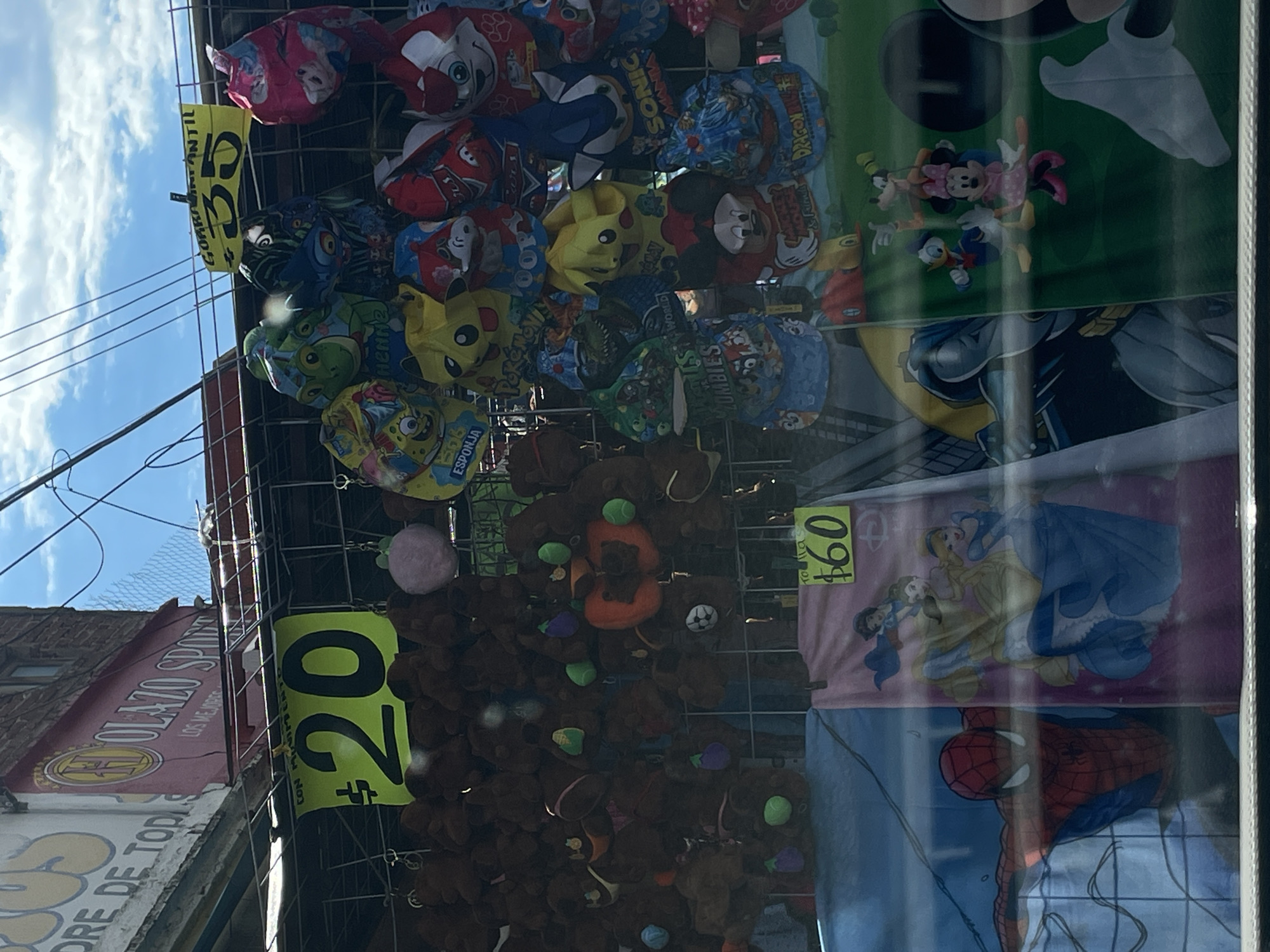 This image shows a storefront display, likely in Oaxaca City, Mexico, featuring a variety of items for sale. The items include character-themed hats featuring designs like Mickey Mouse, Pikachu, and Spiderman, priced at 35 Mexican pesos. There are also plush toys, possibly teddy bears, being sold for 20 Mexican pesos each. Additionally, character-themed towels with designs of Disney princesses and Spiderman are priced at 60 Mexican pesos. The storefront has signage in Spanish. The image appears to be taken from inside a store looking out towards the street, with reflections visible on the glass. A building with the sign "OLAZO SPORT" is visible on the left side of the photo.