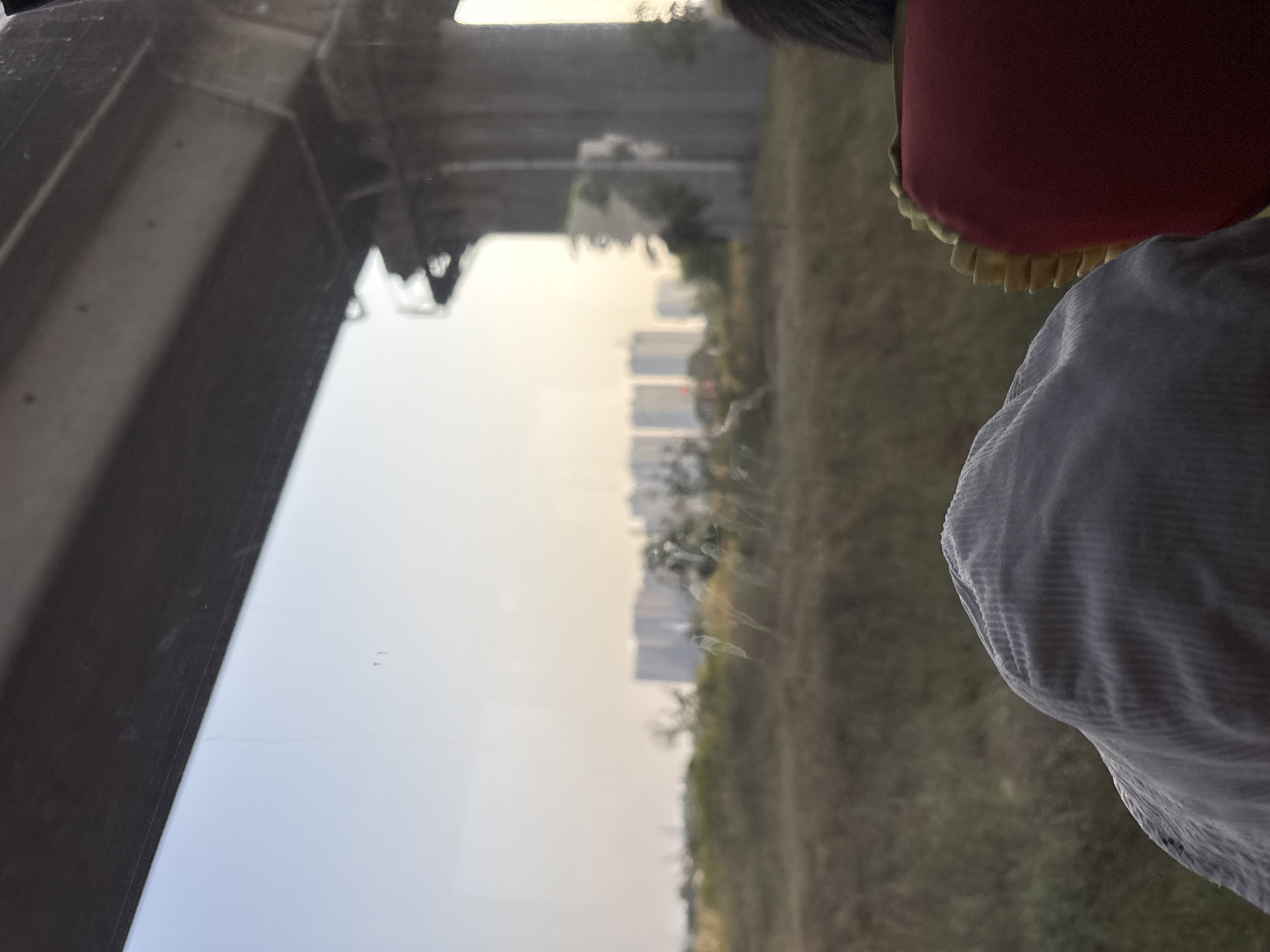 The image captures a view from inside a vehicle looking out a window. In the foreground, the top of a light-colored, corduroy hat is visible, along with the back of a red seat with yellow trim. Beyond the hat and seat, a blurred landscape unfolds, featuring dry grass and vegetation. In the mid-ground, a series of modern buildings, appearing to be apartment complexes, are visible on the horizon. A large concrete structure, possibly part of an overpass or bridge, dominates the right side of the frame. The sky is a pale, hazy yellow, suggesting either sunrise or sunset. The overall scene conveys a sense of travel and observation of the urban fringe.