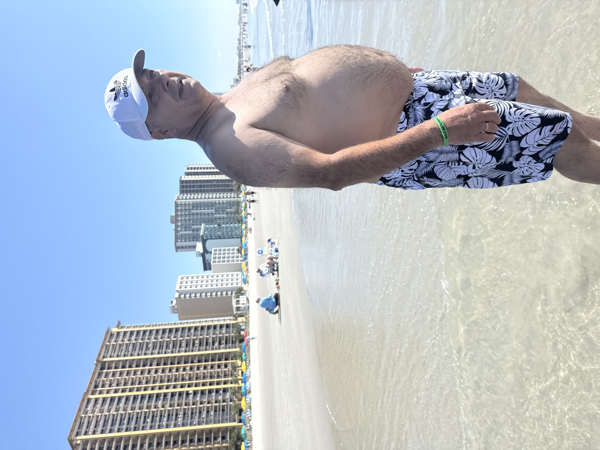 The media file depicts a man standing in shallow water on a beach. He is wearing a white Adidas cap and black and white floral print swim trunks. The man is shirtless, revealing a hairy chest and abdomen. His left arm is extended, and he has a green wristband on it with white text that appears to say "farmhouse.com". He is also wearing a ring on his right ring finger.
In the background, there are several high-rise buildings, suggesting a coastal city location like Myrtle Beach. The sky is clear and blue, indicating a sunny day. Other beachgoers are visible in the distance, some sitting or walking along the shore. Colorful umbrellas are dotted along the beach. The water is clear and shallow, with ripples and reflections on its surface. The scene suggests a relaxed, vacation atmosphere, likely during daytime.