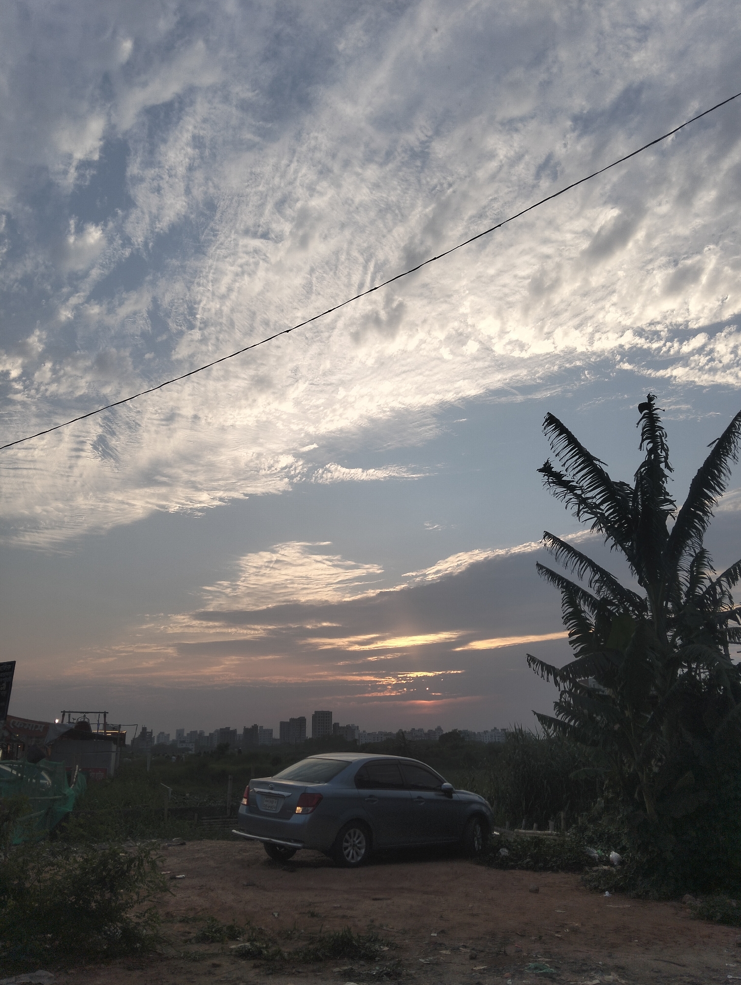 The image captures a gray sedan parked on a dirt lot in Dhaka, Bangladesh. Buildings dot the horizon under a cloudy sky with a hint of sunset. A palm tree stands to the right of the car, and an electrical wire stretches across the upper part of the frame. Vegetation surrounds the parking area.