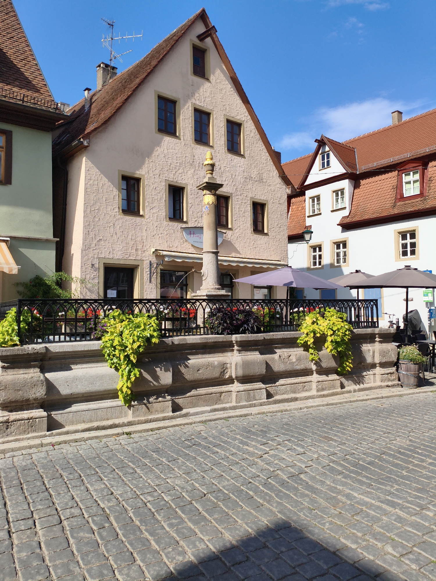 The image depicts the Marktplatz in Rothenburg ob der Tauber, Germany. In the foreground, a cobblestone street leads towards a stone fountain with a decorative column topped by a golden figure. The fountain is surrounded by a black wrought-iron fence, behind which are planters filled with vibrant green ivy and colorful flowers. Beyond the fountain, traditional German architecture dominates the scene. Buildings with steep, red-tiled roofs and stucco facades in shades of cream and pink line the square. Several windows with dark wooden frames are visible on the buildings. Two large umbrellas, one purple and one grey, are set up near tables and chairs, suggesting an outdoor cafe or restaurant. The sky is a clear, bright blue with a few wispy clouds. The sunlight casts distinct shadows across the cobblestone street, indicating a bright, sunny day. There are no people visible in the image.