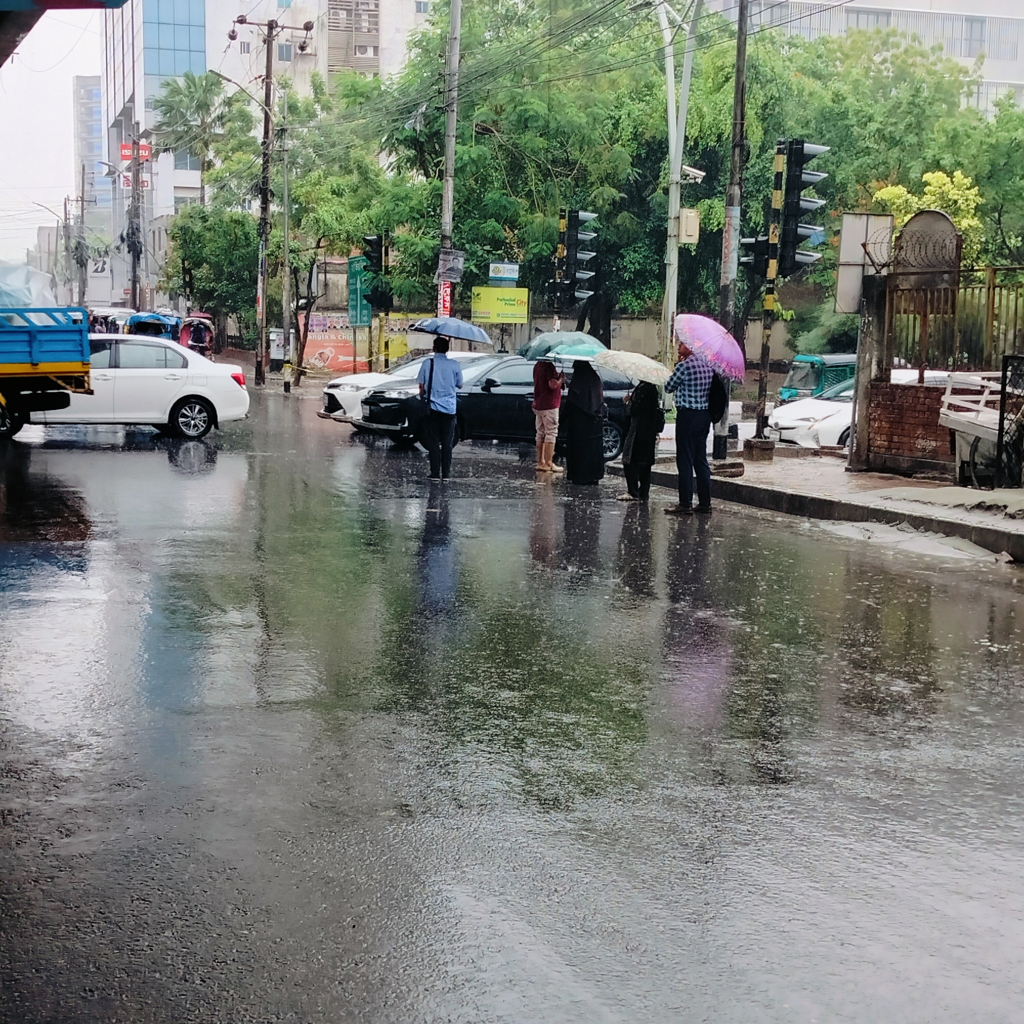 The image depicts an urban street scene in Dhaka, Bangladesh, during or immediately after a rain shower. The asphalt road is thoroughly wet and highly reflective, mirroring the surrounding buildings, trees, and people, suggesting active rain or recent heavy precipitation.
In the foreground, several people are standing near the edge of the wet road, many holding umbrellas of various colors including blue, pink, green, and white/beige. One man in a light blue shirt holds a blue umbrella, while another in a plaid shirt holds a pink umbrella. A woman in a black abaya is also visible under an umbrella. Their reflections are clearly visible on the glistening road surface.
Vehicles are present on the street; a white sedan is seen on the left, and the front of a white car and a black sedan are visible further down. Part of a blue truck bed with a yellow frame is also in the foreground on the left. The scene is bustling with typical urban infrastructure, including multiple utility poles heavily laden with electrical and communication wires, traffic lights, and commercial buildings.
In the background, multi-story buildings line the street, interspersed with lush green trees. Visible text includes "ISUZU" and a partially visible "Bridgestone" logo on building facades. A yellow sign for "Purbochhol Prime City" is also present, along with various posters on utility poles, one mentioning "Bangla & China" and another depicting a "mask up" message. A metal fence topped with barbed wire and a brick wall are seen on the right.
The overall impression is of a busy city street continuing its daily activities despite the wet weather, with people and vehicles navigating the rainy conditions.