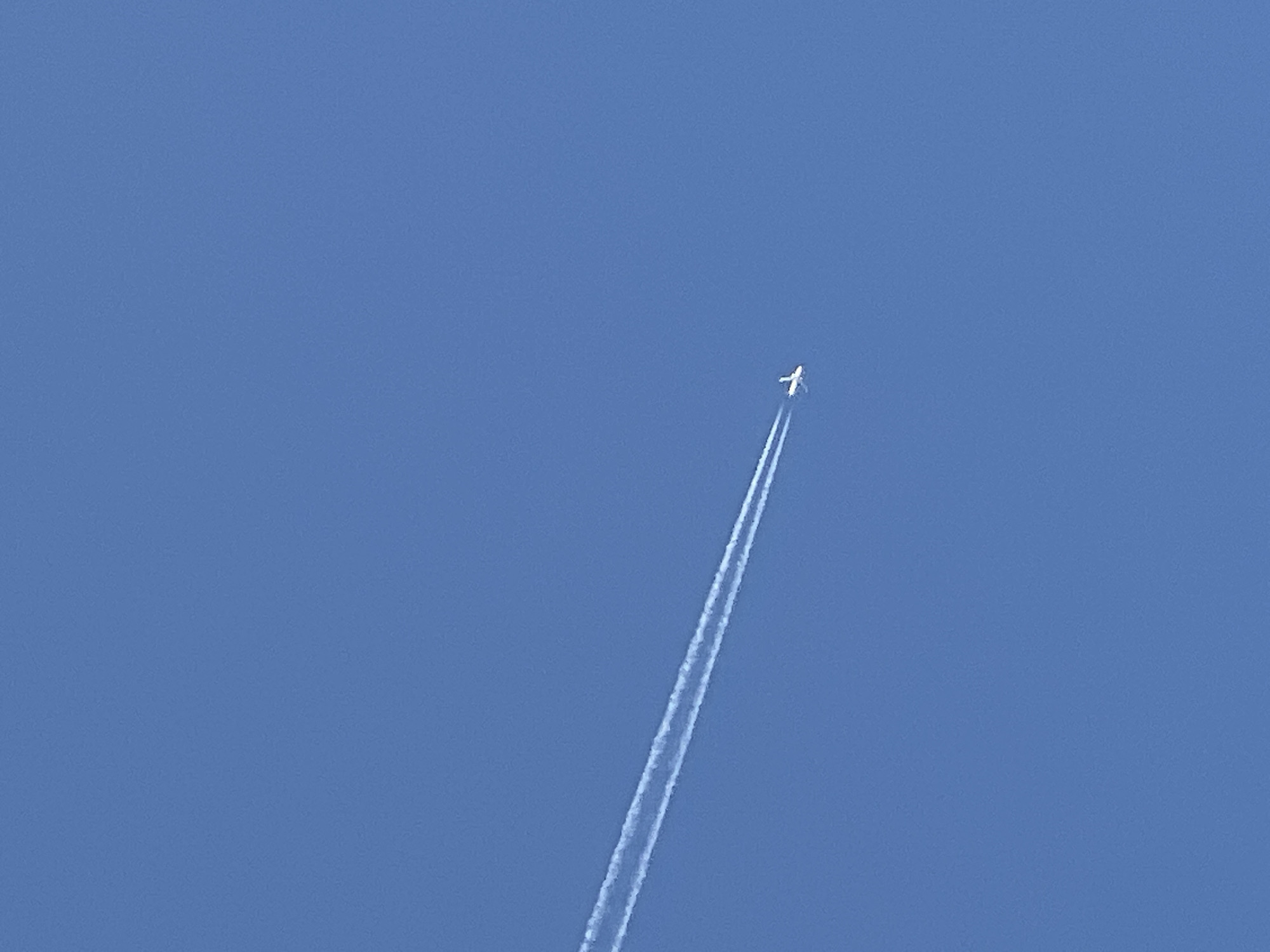 A commercial airplane, identified by its white contrail, is captured in flight against a clear, deep blue sky. The airplane appears as a small white speck with outstretched wings, trailing two parallel lines of white vapor. The contrail stretches diagonally across the frame from the lower left to the upper right. The scene is devoid of any urban or natural landscape features, offering only the vast expanse of the sky. The image was taken in Lublin, Poland.