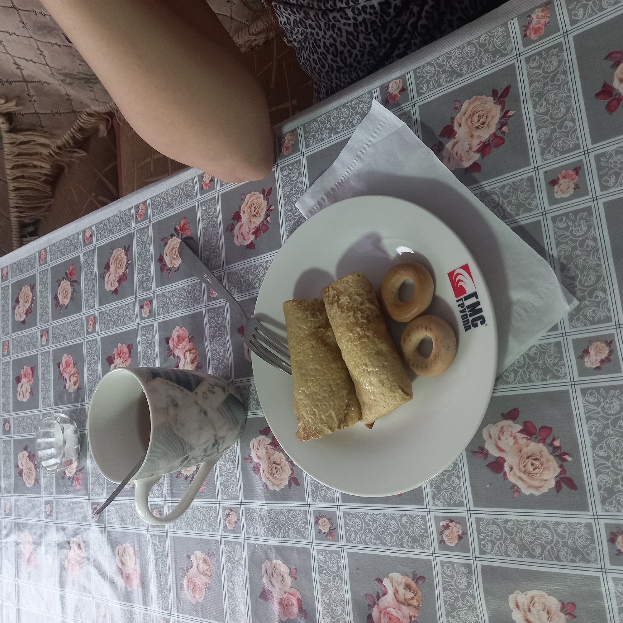 The image depicts a close-up, overhead view of a table setting. The table is covered with a grey tablecloth featuring a repeating pattern of roses and decorative borders. On the table, a white mug with a marble-like pattern contains a dark liquid, likely tea, and a silver spoon. Beside the mug, a small, clear glass bowl holds a white granular substance, possibly sugar.
In the foreground, a white plate holds two golden-brown rolled crepes or blinis, and two small, ring-shaped pastries, possibly bagels or pretzels. The plate has a logo with the letters "TMC" and Cyrillic text below it. A silver fork rests on the plate. A white paper napkin is partially visible beneath the plate.
To the right of the table, a person's bare leg and thigh are visible, with skin tone suggesting daylight exposure. Part of a patterned fabric, possibly upholstery from a couch or chair, is visible in the background, featuring a fringe detail along the top. The lighting suggests an indoor setting with ambient light. The scene is set in Cepeleuți, Moldova, implying a domestic or casual dining environment.