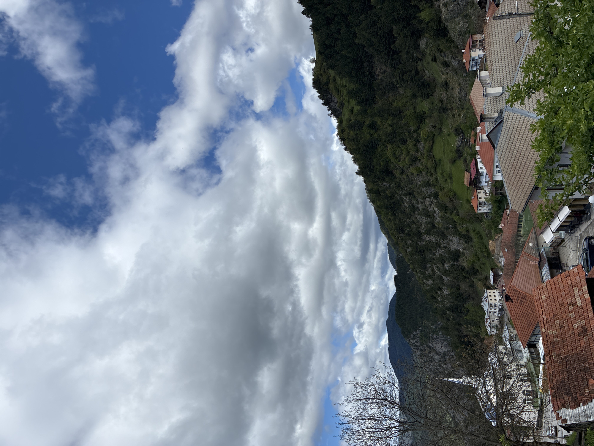 The image shows a view in Giovren, Bulgaria, looking towards a mountainside. The sky is partially cloudy with patches of blue visible. A steep, tree-covered mountainside rises to the right of the frame, with buildings of the town nestled along its base. The buildings have varying roof colors, mostly red and tan. In the foreground are tree branches without leaves.