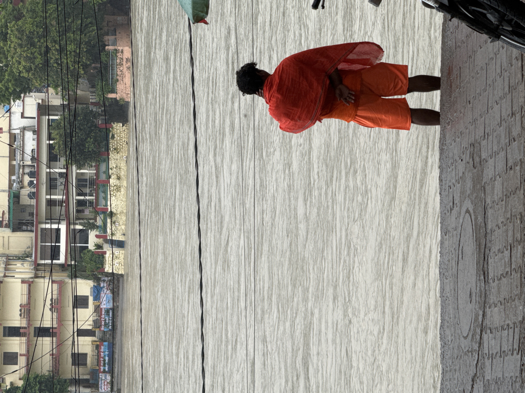 The image depicts a person wearing an orange dhoti and a red shawl, standing in the Ganges River in Rishikesh, India. The person's back is to the camera, and they appear to be looking out at the river. In the background, buildings line the riverbank, with trees and vegetation visible. The river is wide and appears to be flowing strongly. The sky is overcast, suggesting a cloudy or rainy day. The foreground shows a paved walkway or ghat leading down to the river, with a manhole cover visible. The overall scene conveys a sense of peace and spirituality, common in the sacred city of Rishikesh.