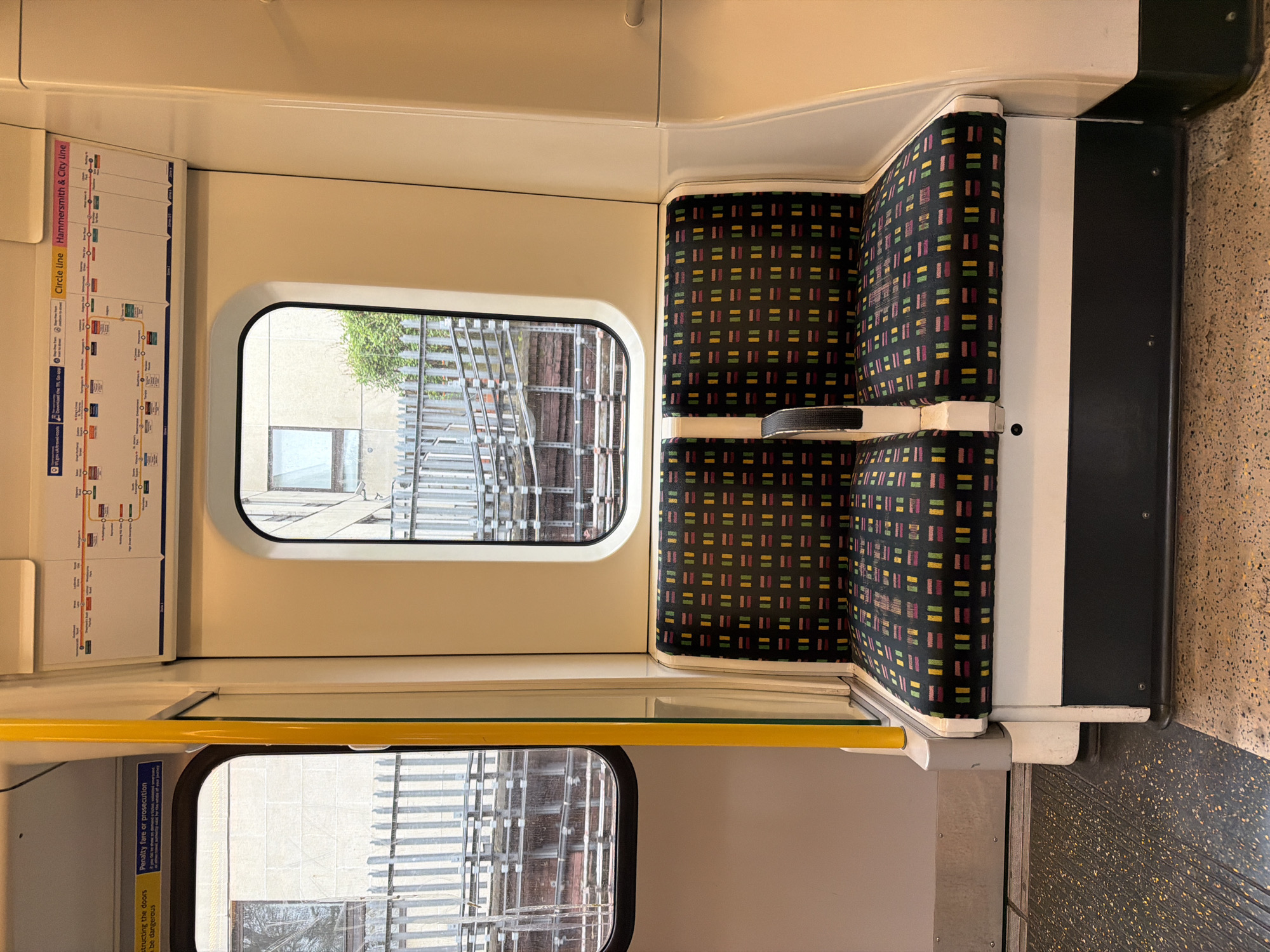 The image shows the interior of a London Underground train carriage. A pair of seats with a distinctive patterned fabric (black background with small colored rectangles) are visible. Adjacent to the seats are windows offering a glimpse outside, showing scaffolding and a building. A yellow handrail runs horizontally. A schematic map of the Circle and Hammersmith & City lines is posted above the window, and a notice about penalties for obstructing the doors is visible below the window. The color palette is muted, with creams, grays, and blacks predominating.