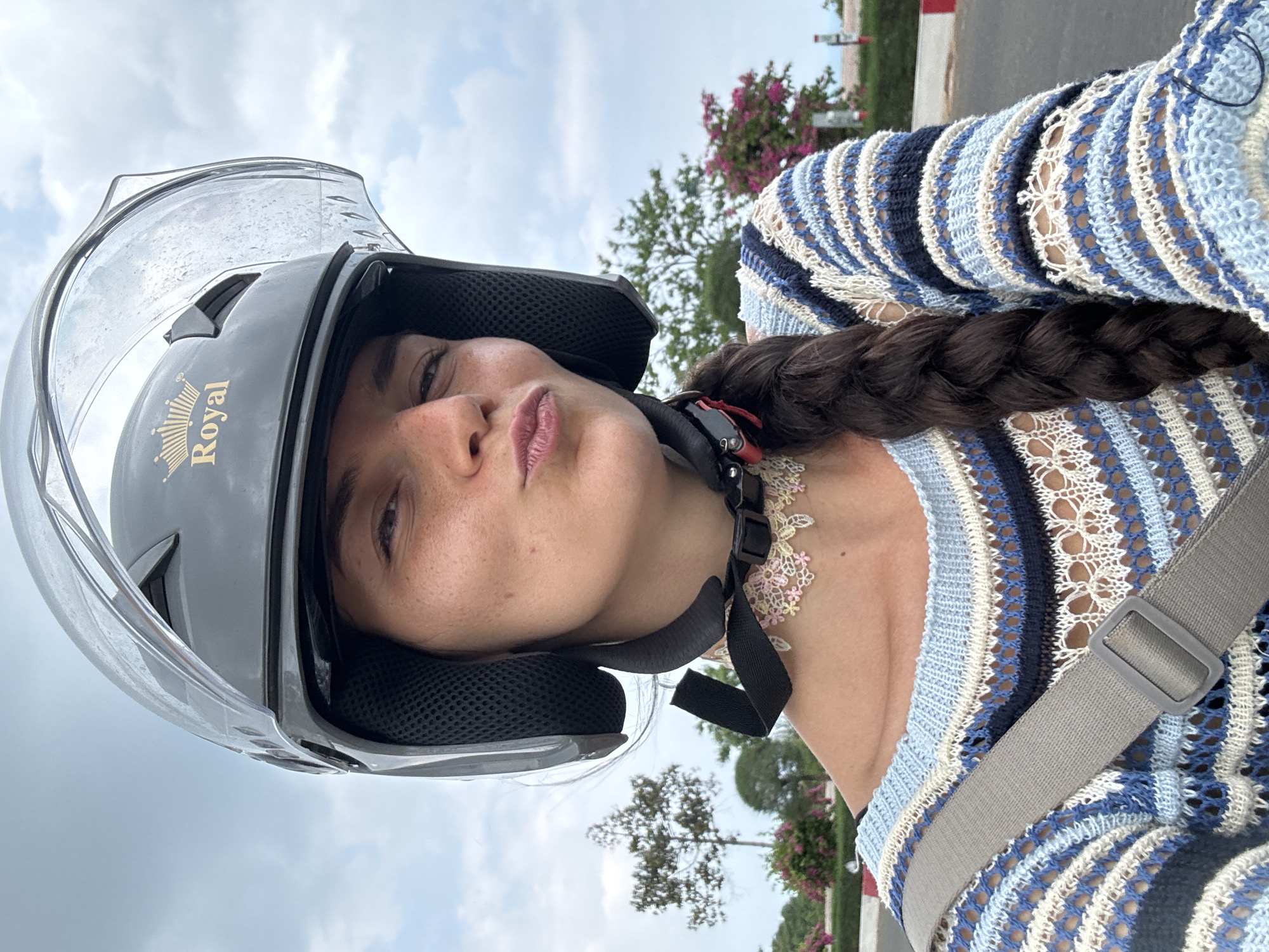 A medium shot captures a young woman wearing a black motorcycle helmet with a clear visor and a crown emblem labeled "Royal" at the front. She is looking directly at the camera with her lips puckered, her face adorned with freckles. Her dark brown hair is styled in a thick braid that falls over her shoulder. She is wearing a light blue, white, and navy striped crochet sweater with a grey strap across her chest. A delicate pink and yellow lace choker is visible around her neck.
The background suggests an outdoor, possibly urban or suburban setting in Vietnam, with hints of trees, pink flowering bushes, and a road with a red and white striped curb. The sky is overcast with grey clouds. The overall impression is a candid selfie taken outdoors, possibly during a scooter ride.