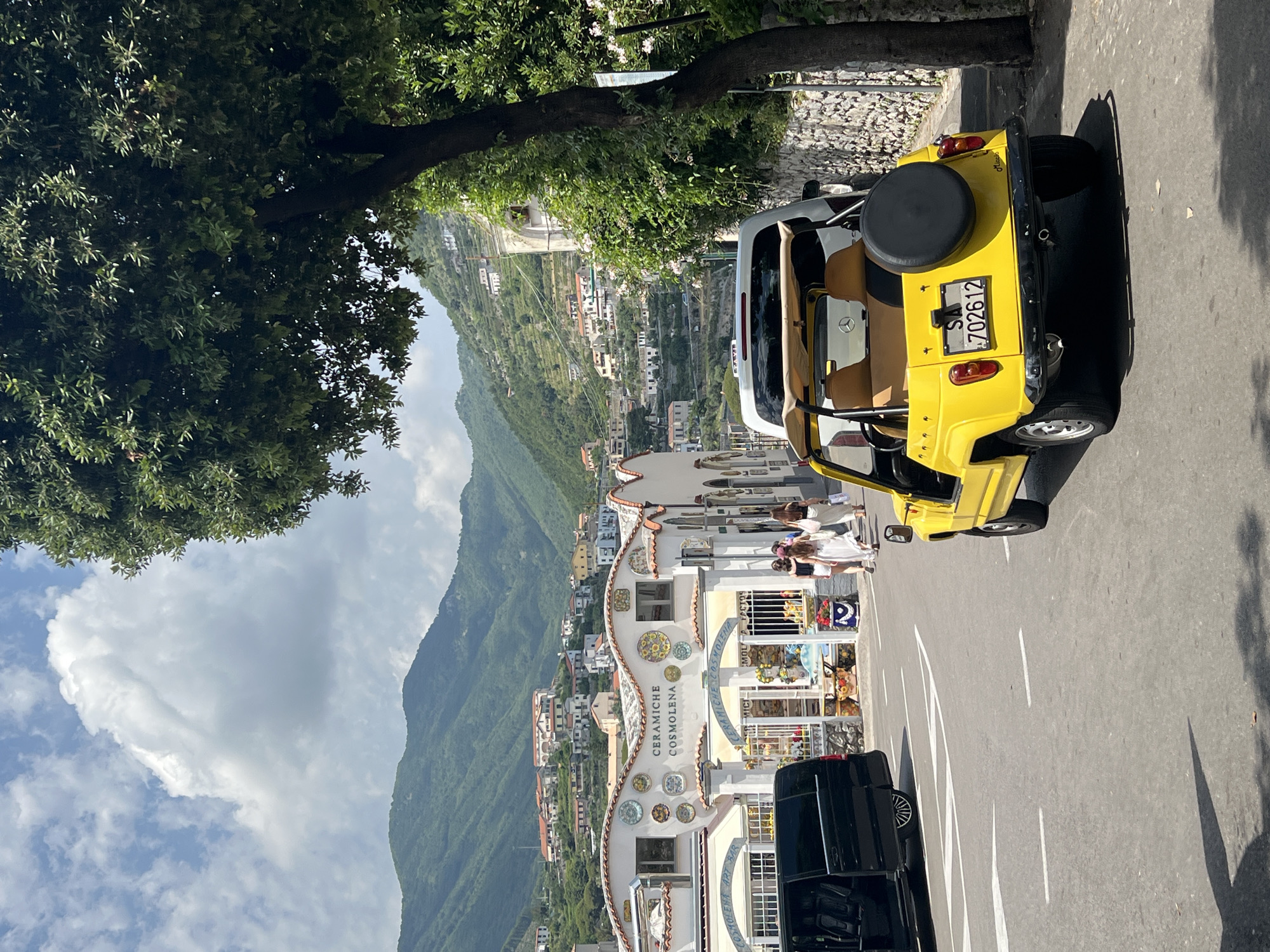 The image captures a street scene in Ravello, Italy, with a vibrant yellow car parked on the right side of the road. The car, a convertible with its top down, sports a spare tire on the back and a license plate reading "SA 702612". In the background, a white SUV is also parked. To the left, a black van with its rear door open is visible, and several pedestrians, appearing to be women dressed in light-colored clothing, are walking away from the camera.
The buildings lining the street are white with decorative ceramic plates on their facades. One building prominently displays the text "CERAMICHE COSMOLENA". The street itself is paved and marked with white lines. Behind the buildings, the lush green hills of Ravello rise, dotted with more white houses and buildings cascading down the slopes. The sky is blue with fluffy white clouds, and a large tree with dense green foliage is in the foreground on the right, partially obscuring the view of the hills. The overall impression is a sunny day in a picturesque Italian town.