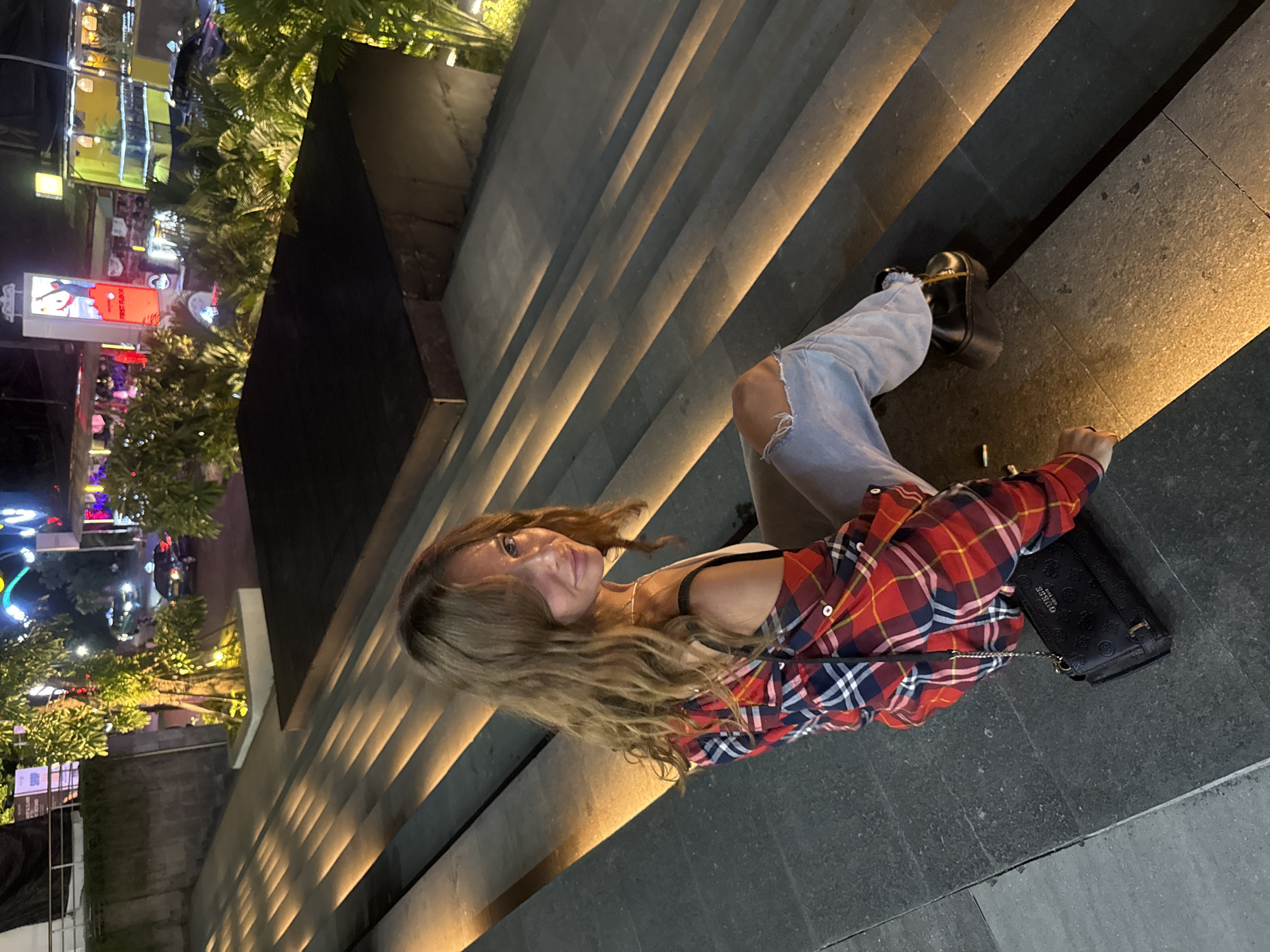 A woman with light brown hair is sitting on a set of gray stone steps illuminated with warm-toned recessed lighting. She is wearing ripped blue jeans, a white tank top, and a red and blue plaid shirt. A black purse rests near her on the steps. The steps appear to be part of a larger structure, potentially a building or raised platform.
In the background, city lights and illuminated signs are visible. Vegetation is also present, suggesting an urban environment with landscaping. The location is Kuta, Indonesia, based on the visual characteristics of the architecture, vegetation, and city lights.