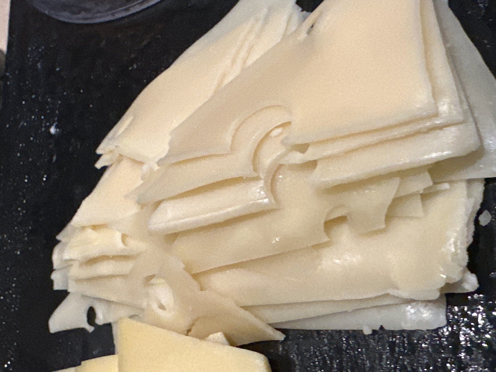The image presents a close-up, high-angle view of a stack of thinly sliced, light yellow food items, resembling cheese, arranged on a dark, reflective surface. The slices are irregularly shaped, with many exhibiting distinct, rounded perforations or holes, similar to Emmental or Swiss cheese. The texture appears smooth with a slight sheen. A smaller cluster of uniformly shaped, darker yellow slices is visible on the far left. The background consists of a black or very dark platter, showing subtle reflections and sparse, faint white speckles. A dark, circular edge, possibly part of another serving dish, is partially visible on the upper right. No individuals, specific actions, or direct environmental indicators are present. The scene is depicted as taking place in the city of Púng Luông, Vietnam.