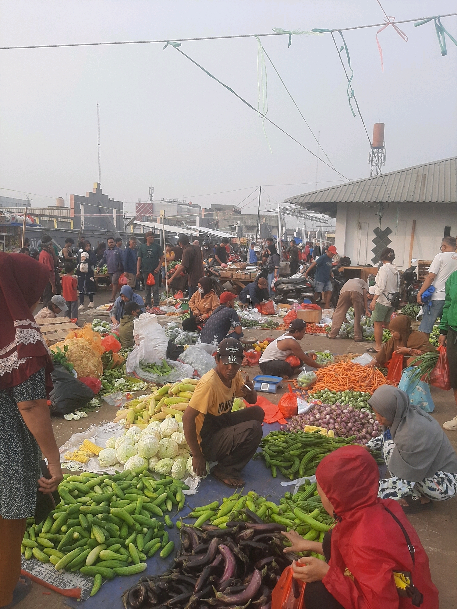This image depicts a bustling outdoor market scene in Bekasi, Indonesia. A variety of fresh produce, including cucumbers, cabbage, carrots, onions, and eggplants, are displayed on the ground, some arranged on tarps and others in plastic bags. Numerous individuals, both men and women, are seen engaged in various activities such as selling, browsing, and interacting with each other. Many are dressed in casual attire, with some women wearing hijabs. The market appears to be set up on a street or open area with buildings and structures visible in the background. The sky is hazy, suggesting it might be early morning or a day with pollution. A tower with an antenna is also visible in the distance. The overall atmosphere suggests a vibrant, local marketplace environment.
