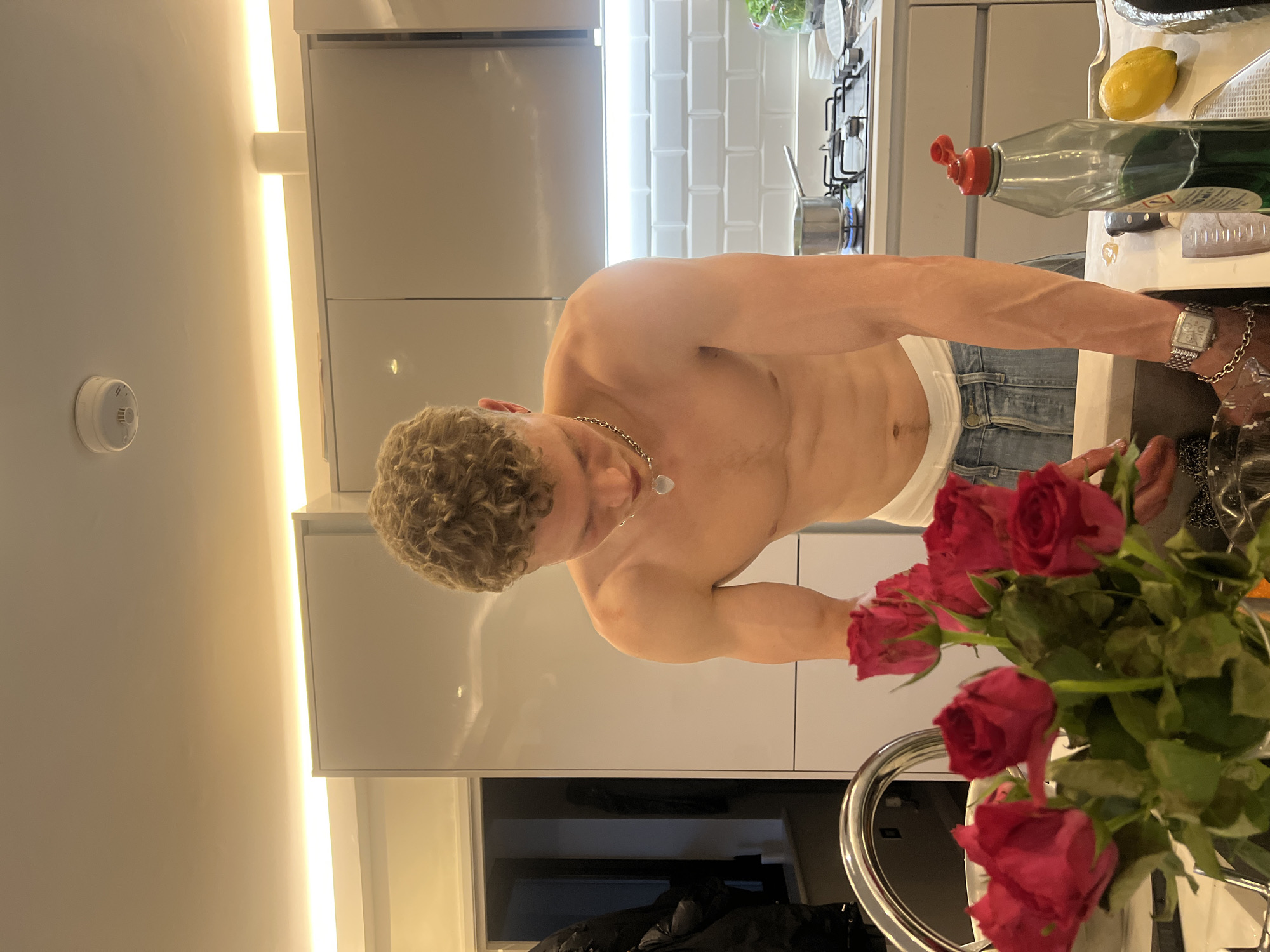 The image captures a shirtless man engaged in an activity at a kitchen sink in what appears to be a modern kitchen. The man, with curly light brown hair, wears light blue jeans with a white undershirt band visible above them. A silver chain with a pendant adorns his neck, and a silver watch is on his left wrist. He is focused on the task at hand, with his hands near or in the sink area on the right side of the frame.
The kitchen features sleek, light grey or white glossy cabinets. Behind the man, a white tiled backsplash and a gas hob with black grates and a silver saucepan are visible. On the counter next to the sink, a green bottle of dish soap with a red cap, a yellow lemon, and several knives can be seen.
In the immediate foreground, to the bottom right, a bouquet of red roses in a silver vase partially obscures the view. The overall lighting is bright, with a vertical LED strip light running along the wall or cabinet edge on the left, contributing to a well-lit indoor environment. A white circular smoke detector is affixed to the wall in the upper left portion of the image. The setting is a contemporary kitchen, likely in a residential building in London, United Kingdom. No discernible text is visible on any objects.