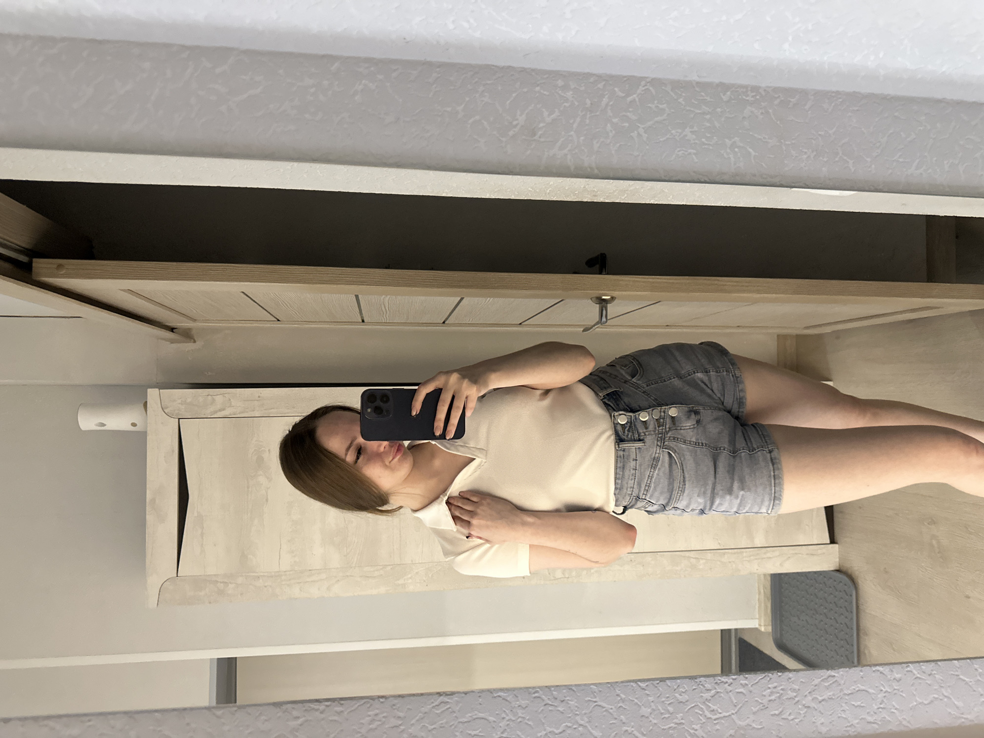 A mirror selfie captures a woman in a bathroom setting. She is wearing a white collared shirt and gray denim shorts. The bathroom features neutral-toned cabinetry, a toilet paper holder, and patterned wall coverings.